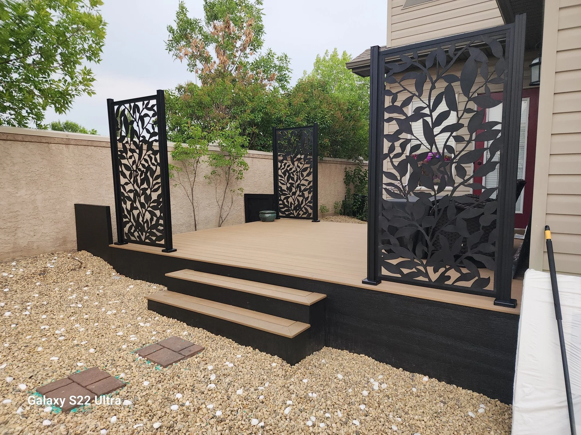 A backyard patio with a wooden deck, decorative black panels with leaf patterns, a gravel ground, a beige wall, with trees and a house in the background.