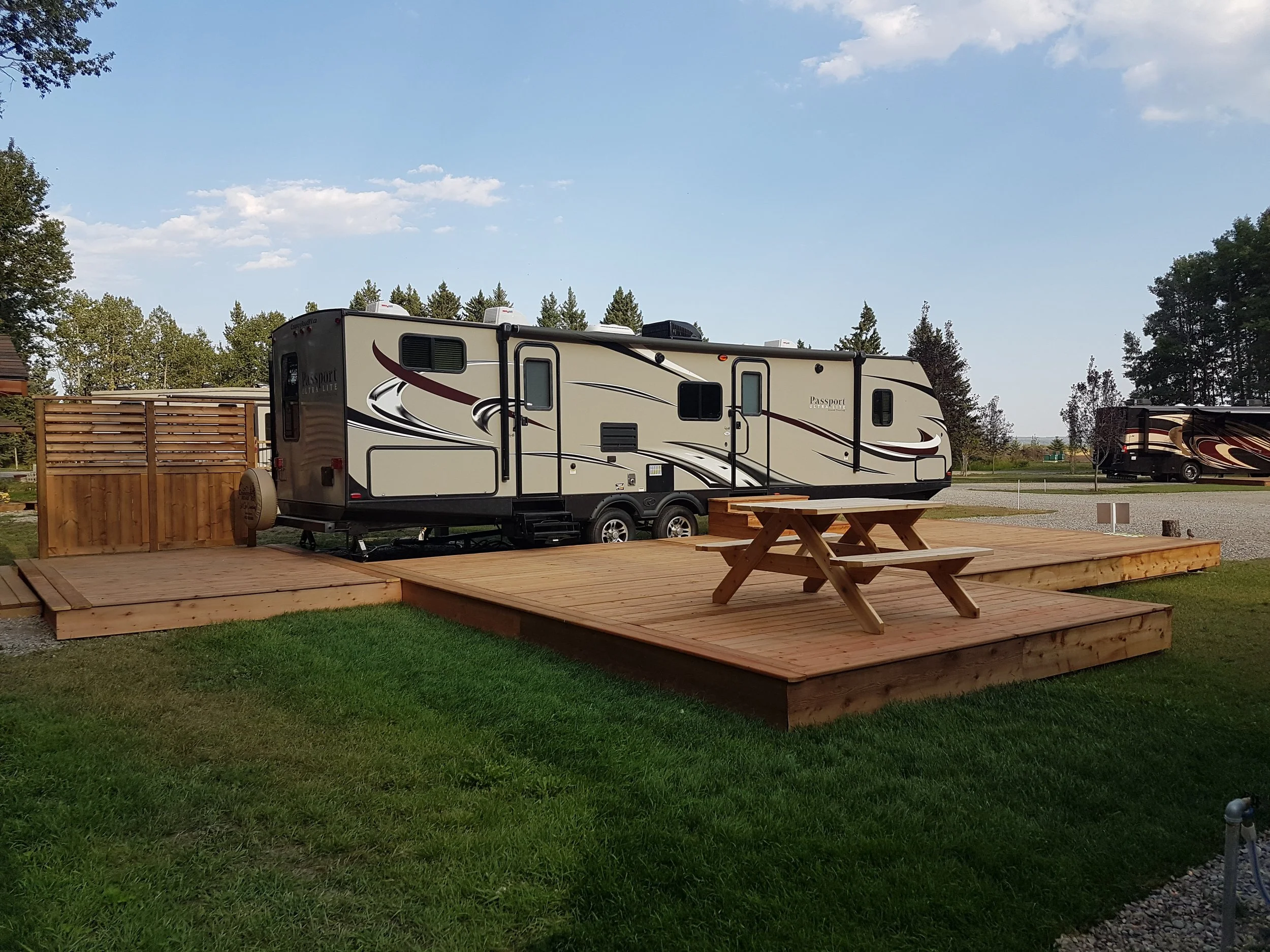 A recreational vehicle (RV) parked on a wooden platform with a picnic table nearby. The RV is beige with black and maroon accents. There is a wooden fence on the left side of the image, and trees and a partly cloudy sky in the background.