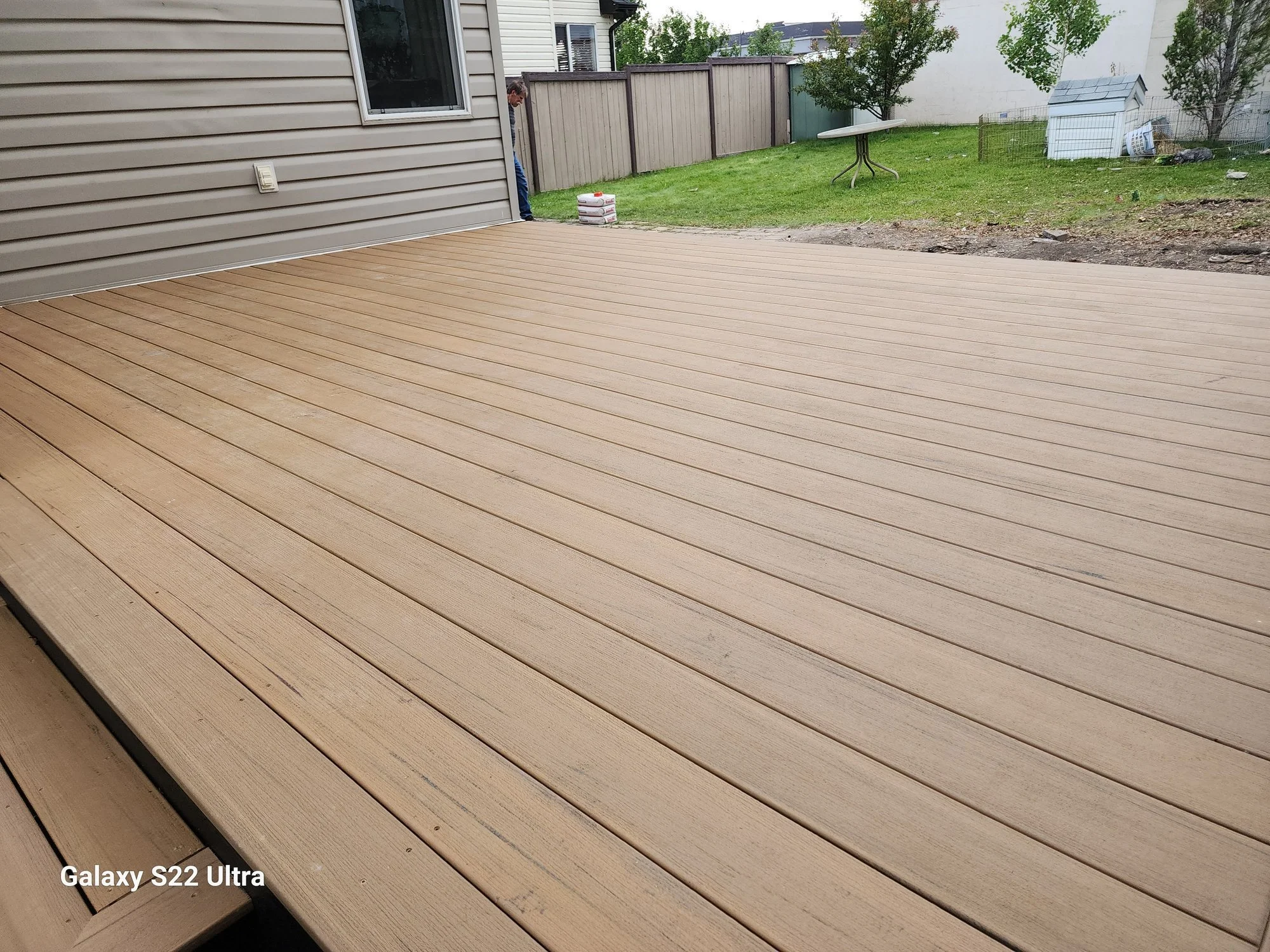 A backyard patio with beige composite decking, a house wall with beige siding, a window, a man looking down at a stack of cement bags, a grassy area with trees, a round folding table, a dog house, and garden fencing.