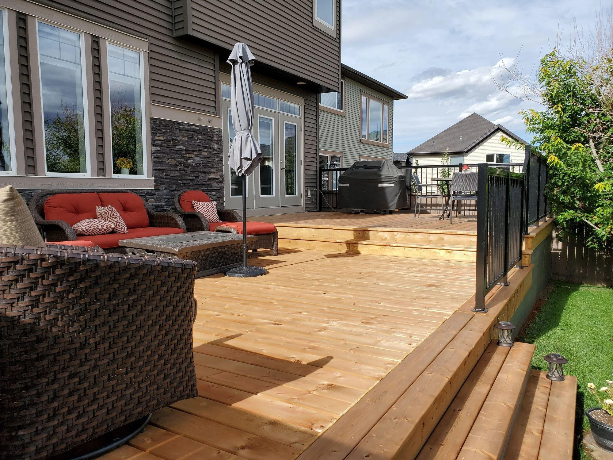 A wooden deck on the back of a house with outdoor furniture, a barbecue grill, and an umbrella, surrounded by trees and neighboring houses under a partly cloudy sky.