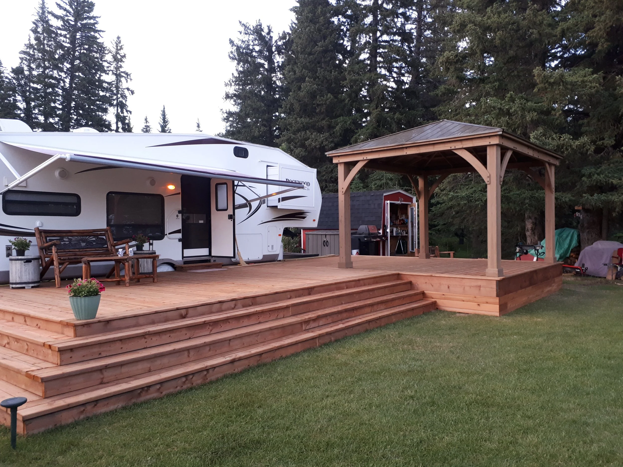 A wooden deck with stairs leading up to a wooded area with tall pine trees, a white RV parked on the deck, a small shed, and outdoor furniture under a wooden gazebo.