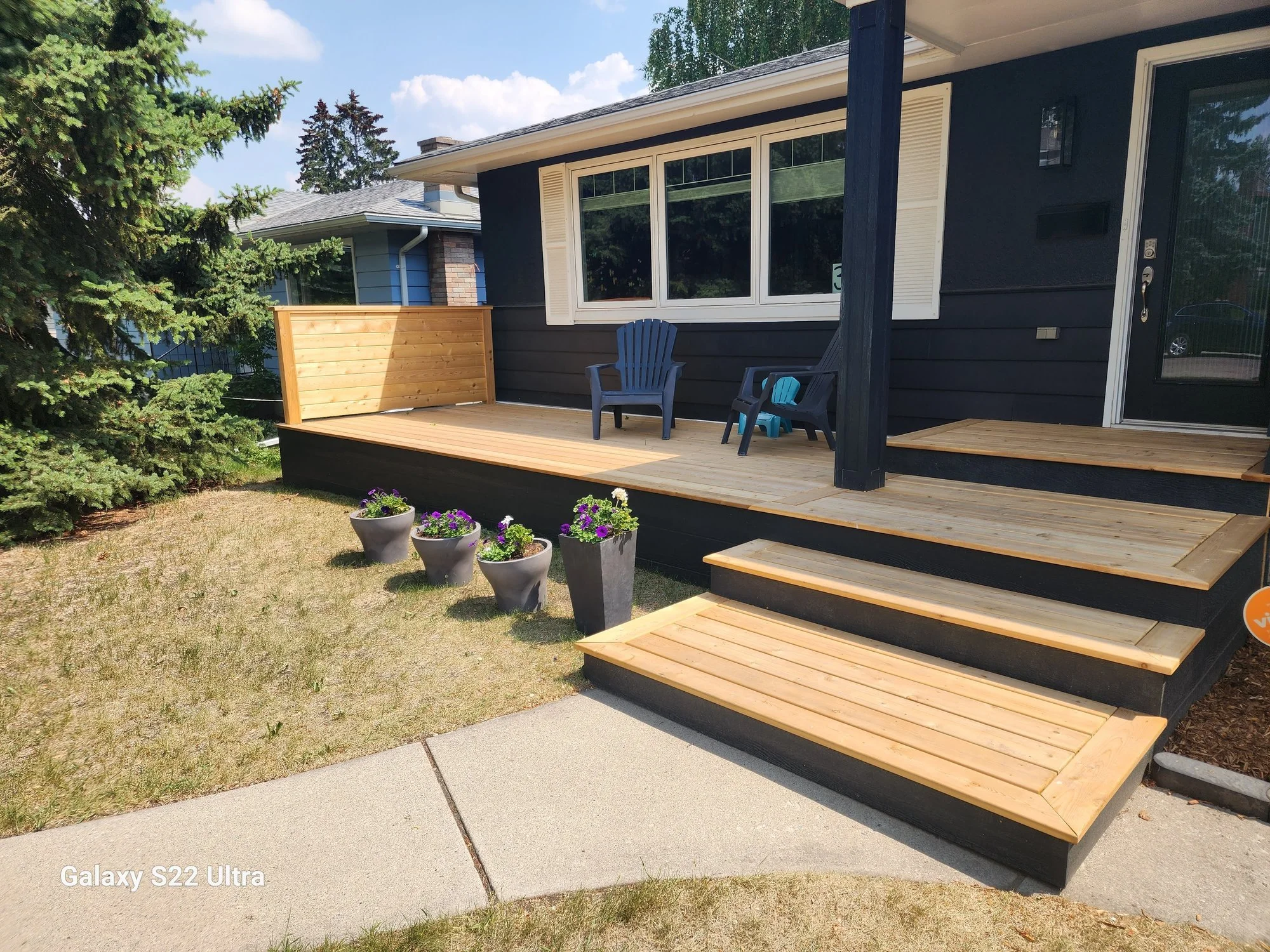 A wooden deck attached to a house with black siding. There are three plastic chairs on the deck, two high-backed dark blue chairs and one small blue chair. Potted flowers are in the foreground, and large trees are in the background. The house has a large window with white shutters and a glass door.