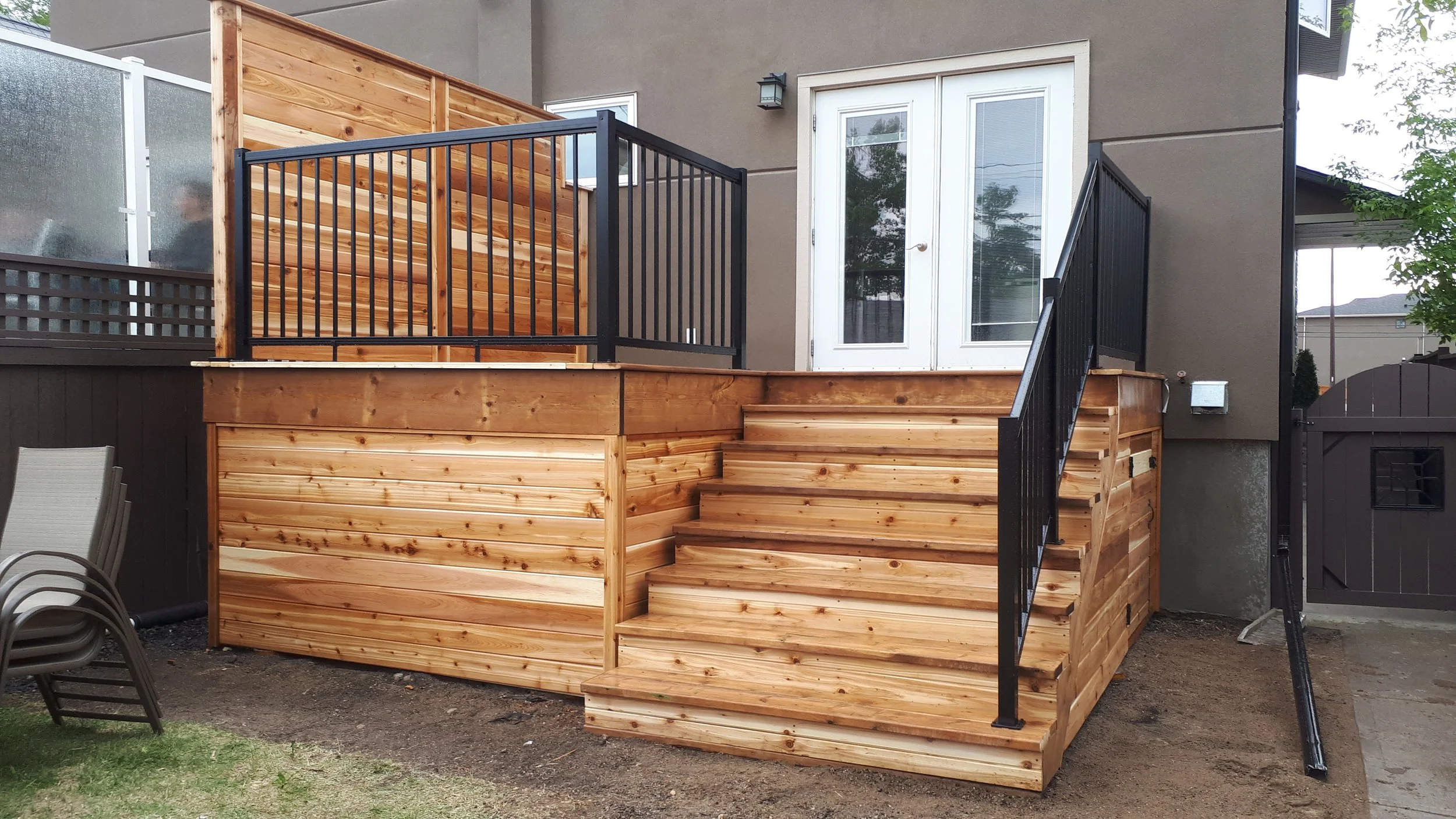A wooden deck with stairs attached to a house, featuring black metal railings, a sliding glass door, and a wooden privacy fence.