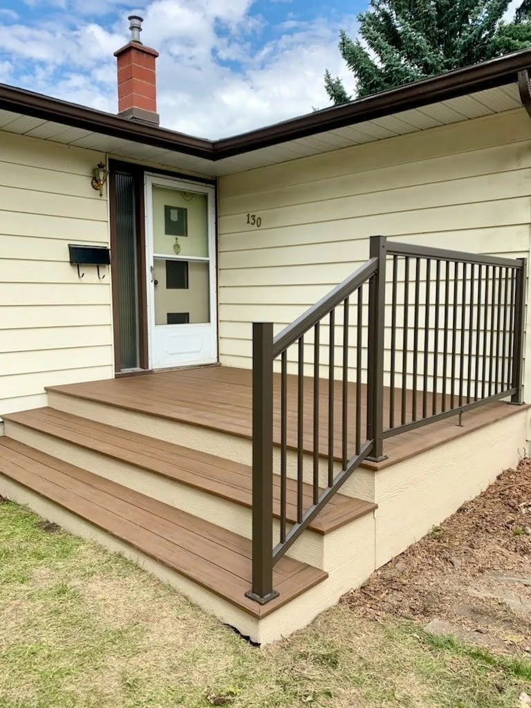 Front porch of a house with light yellow siding, a small wooden deck, black metal railing, and three steps leading to the door, with house number 130 displayed on the wall.