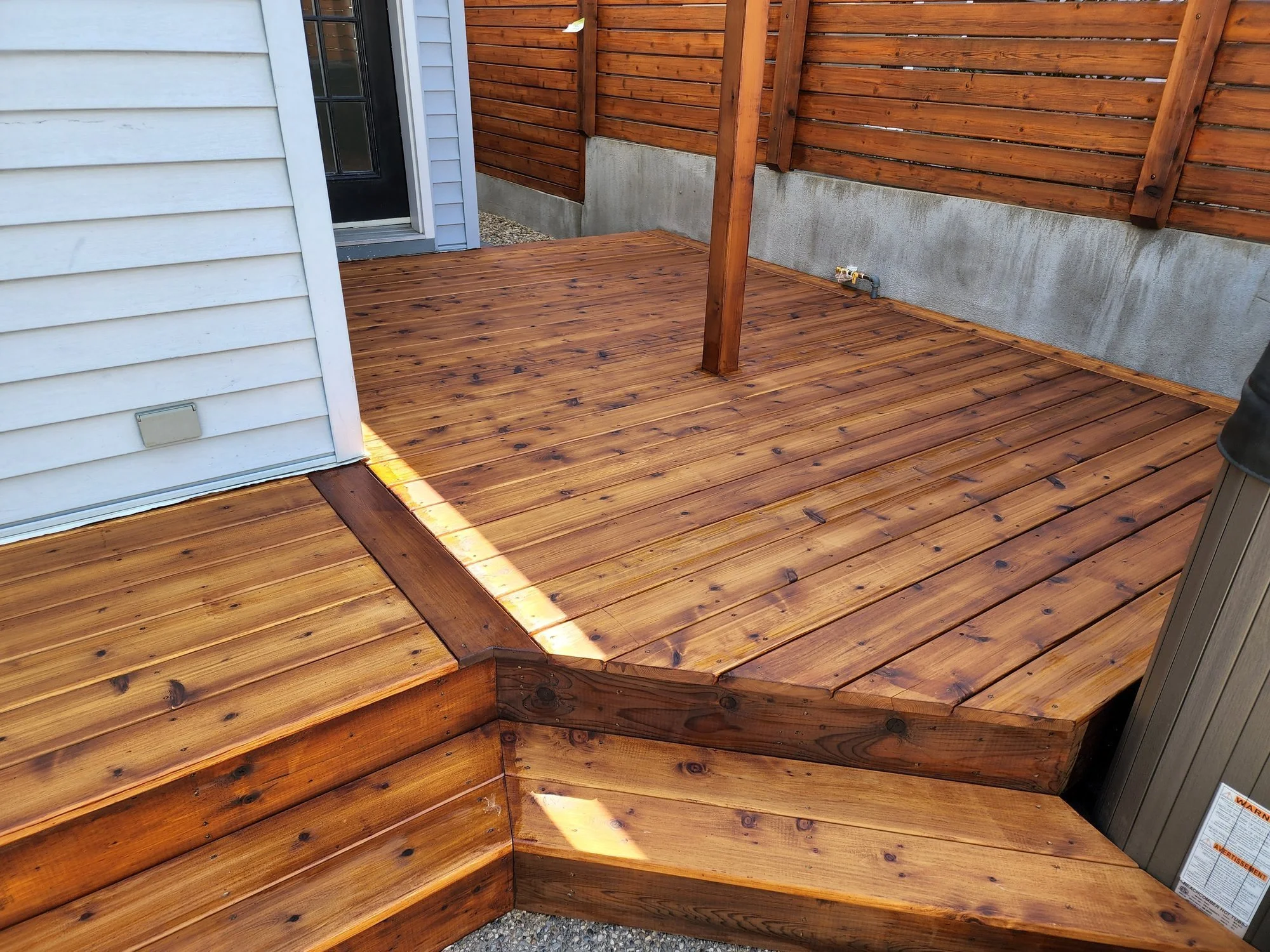 Freshly stained wooden deck with a small step and a corner where the wood meets a concrete foundation, adjacent to a house with light blue siding and a black door.