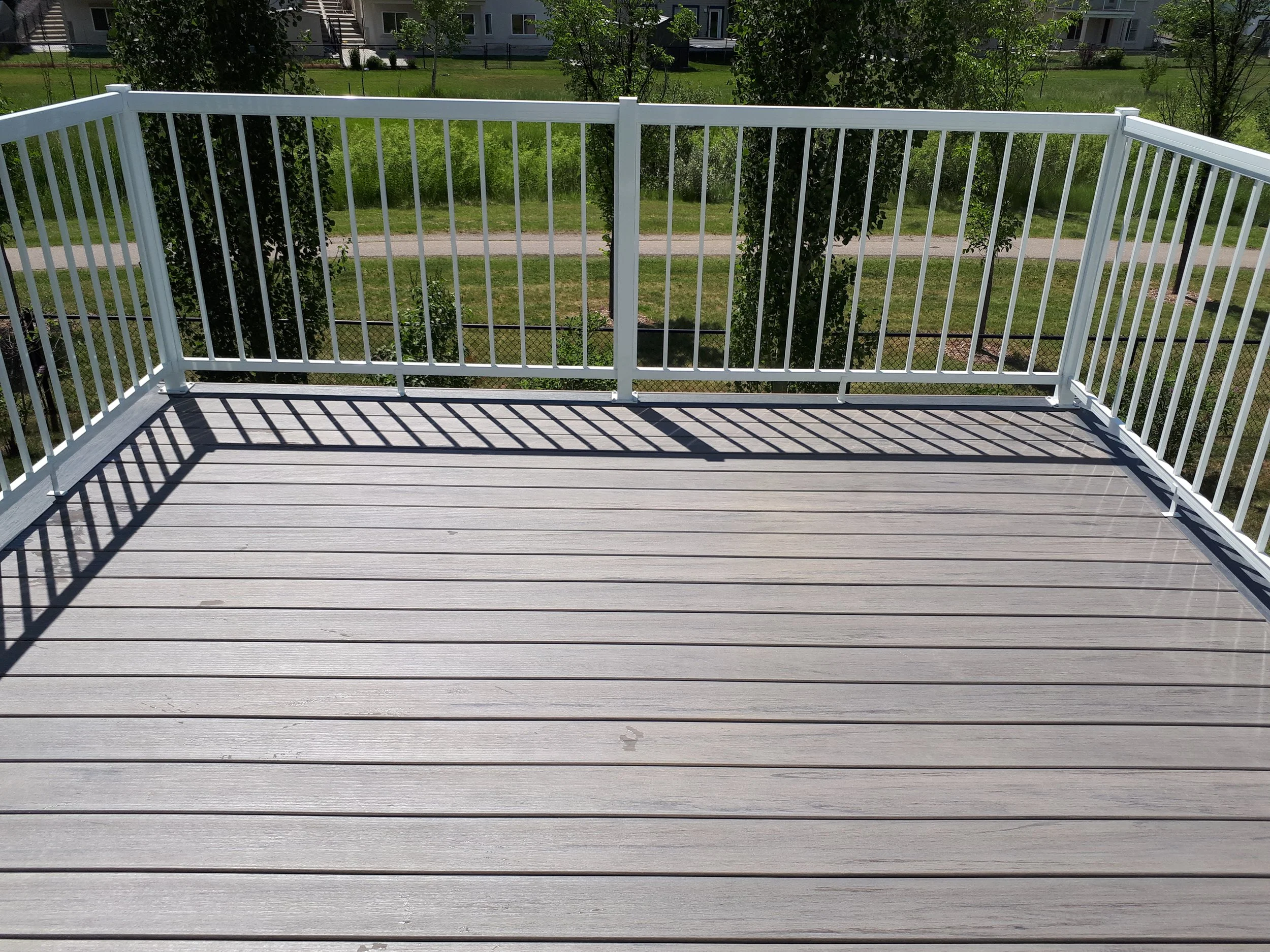 A white balcony railing with shadow lines cast onto the gray decking floor, overlooking a grassy area with trees and buildings in the background.