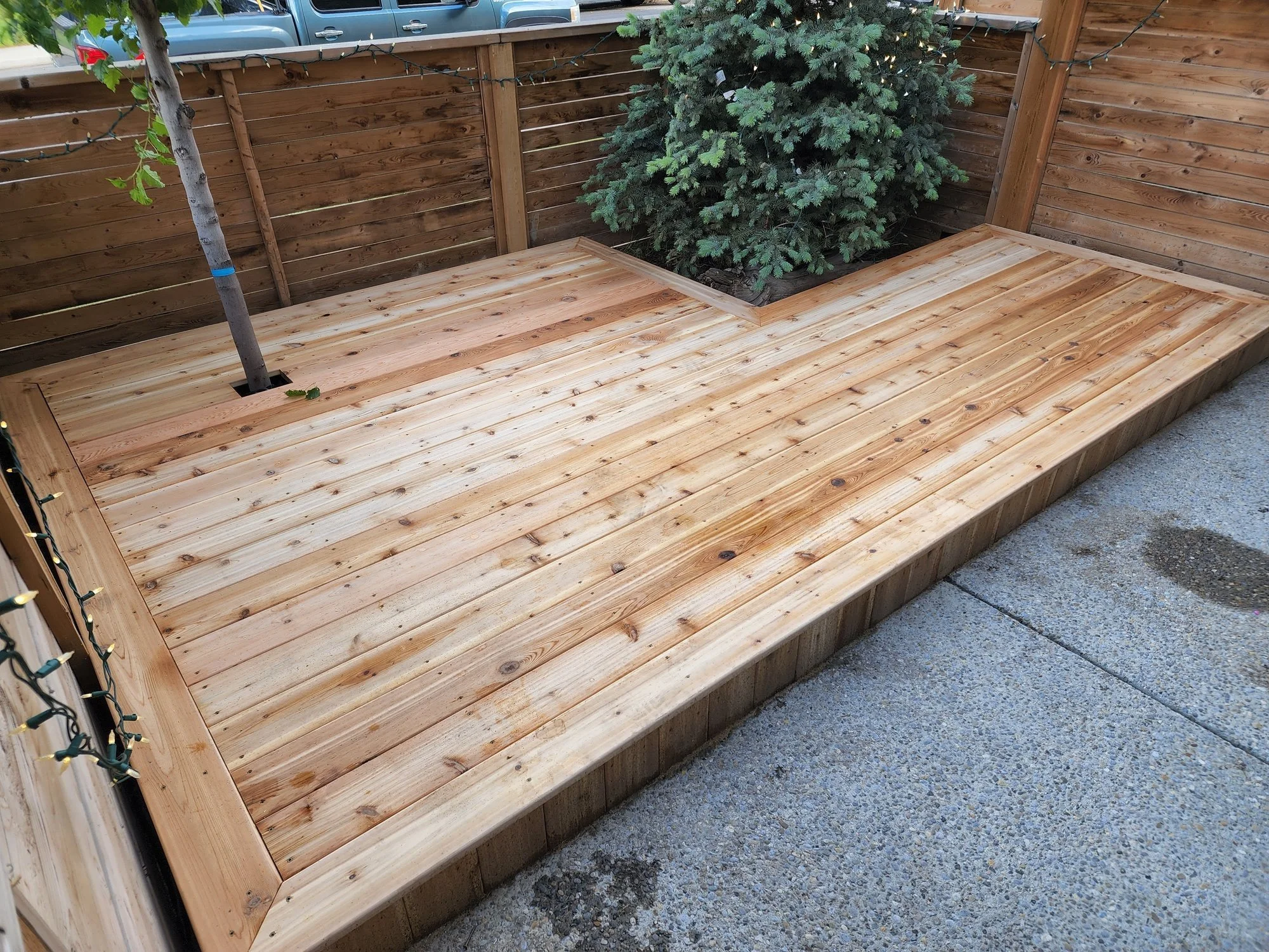 A newly built wooden deck with a built-in planter containing a small evergreen tree, enclosed by a wooden privacy fence, with string lights hanging along the fence and a parked vehicle visible behind it.