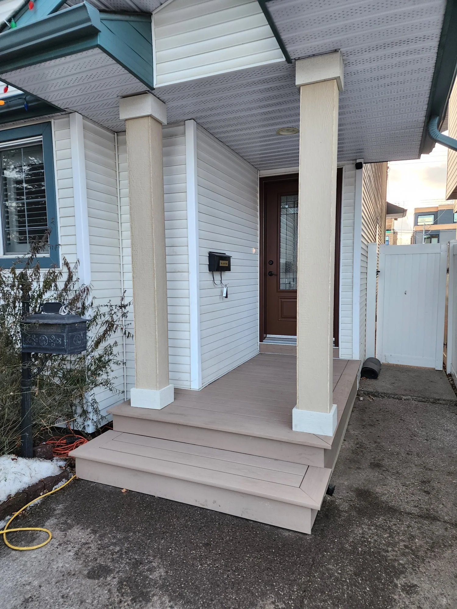Front porch with beige wooden deck and columns, a dark brown front door with glass panel, white vinyl siding, and a white picket fence in the background. There is a mailbox and some plants near the porch.