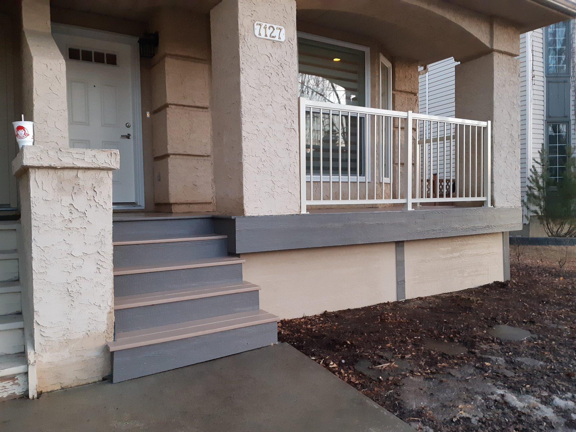 Front porch of a house with a light beige stucco exterior, white front door, and metal railing on a small balcony. The house number '7127' is on a white plaque above the window. There are stairs leading up to the porch, and a large soda cup with a straw sitting on a low wall near the door. The area around the porch has bare soil and some scattered leaves.