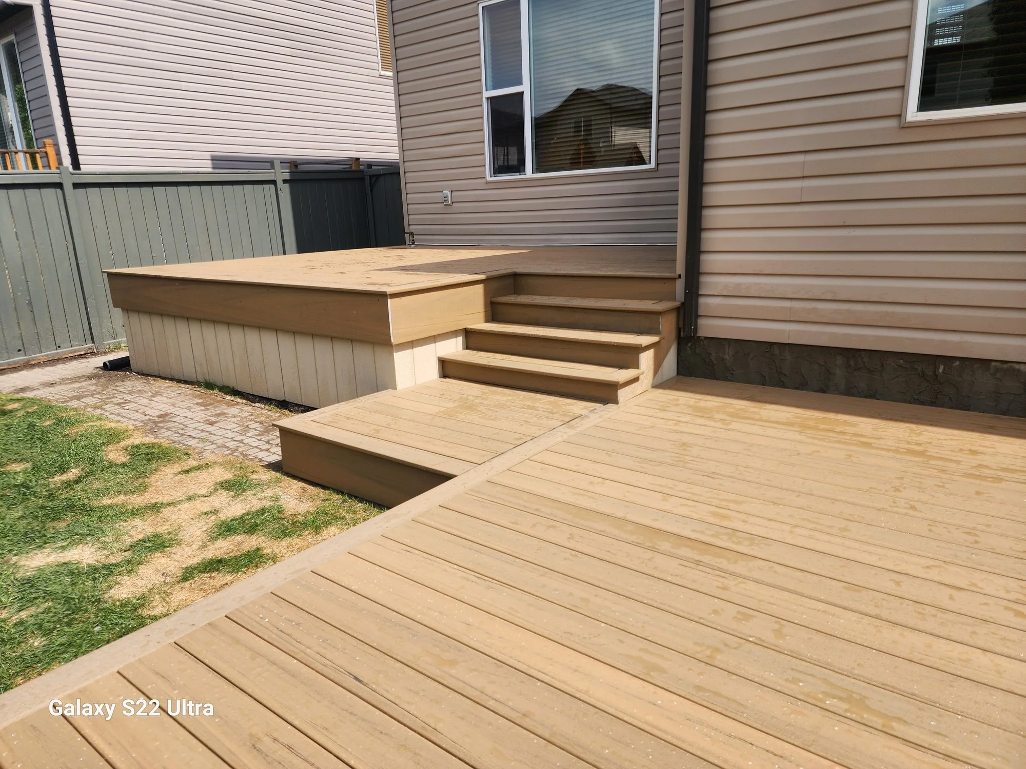 Wooden deck with stairs leading up to the back door of a beige house, fenced yard with grass and brick pathway.