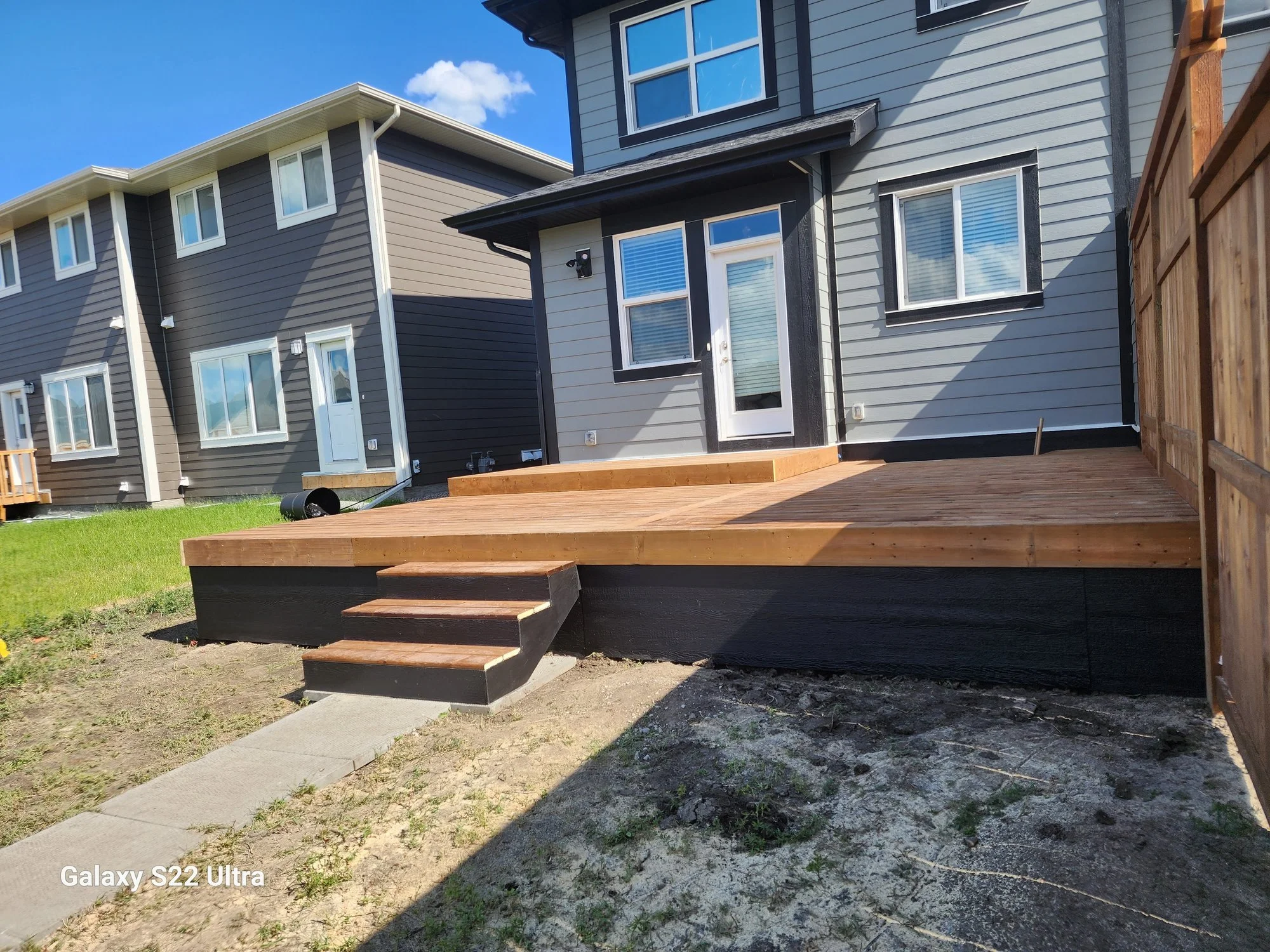 Backyard with wooden deck, stairs leading down to dirt and grass, neighboring townhouses with gray and beige siding, blue sky with clouds.