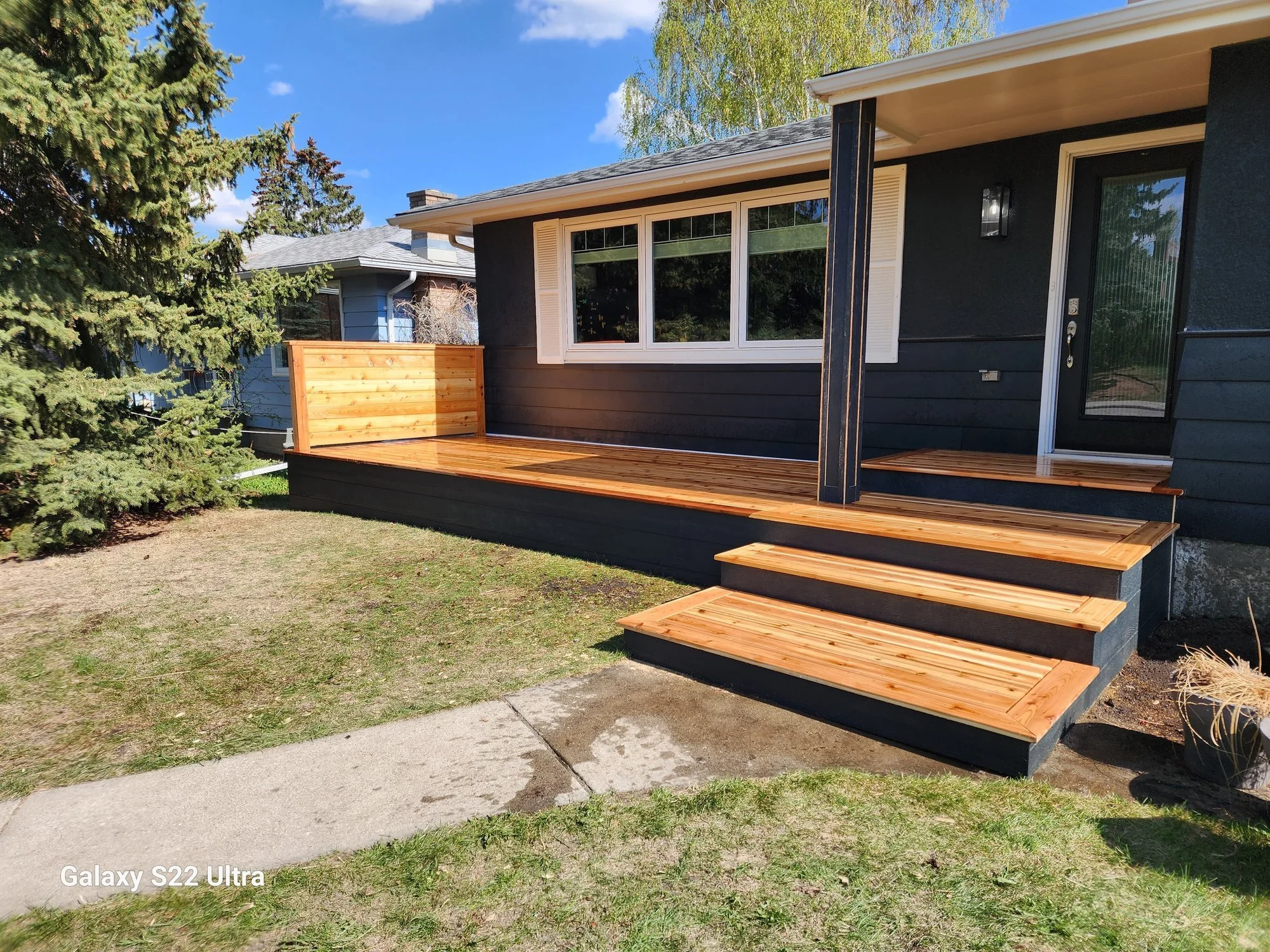 A black house with a wooden deck outside, surrounded by trees and a lawn, under a blue sky with some clouds.