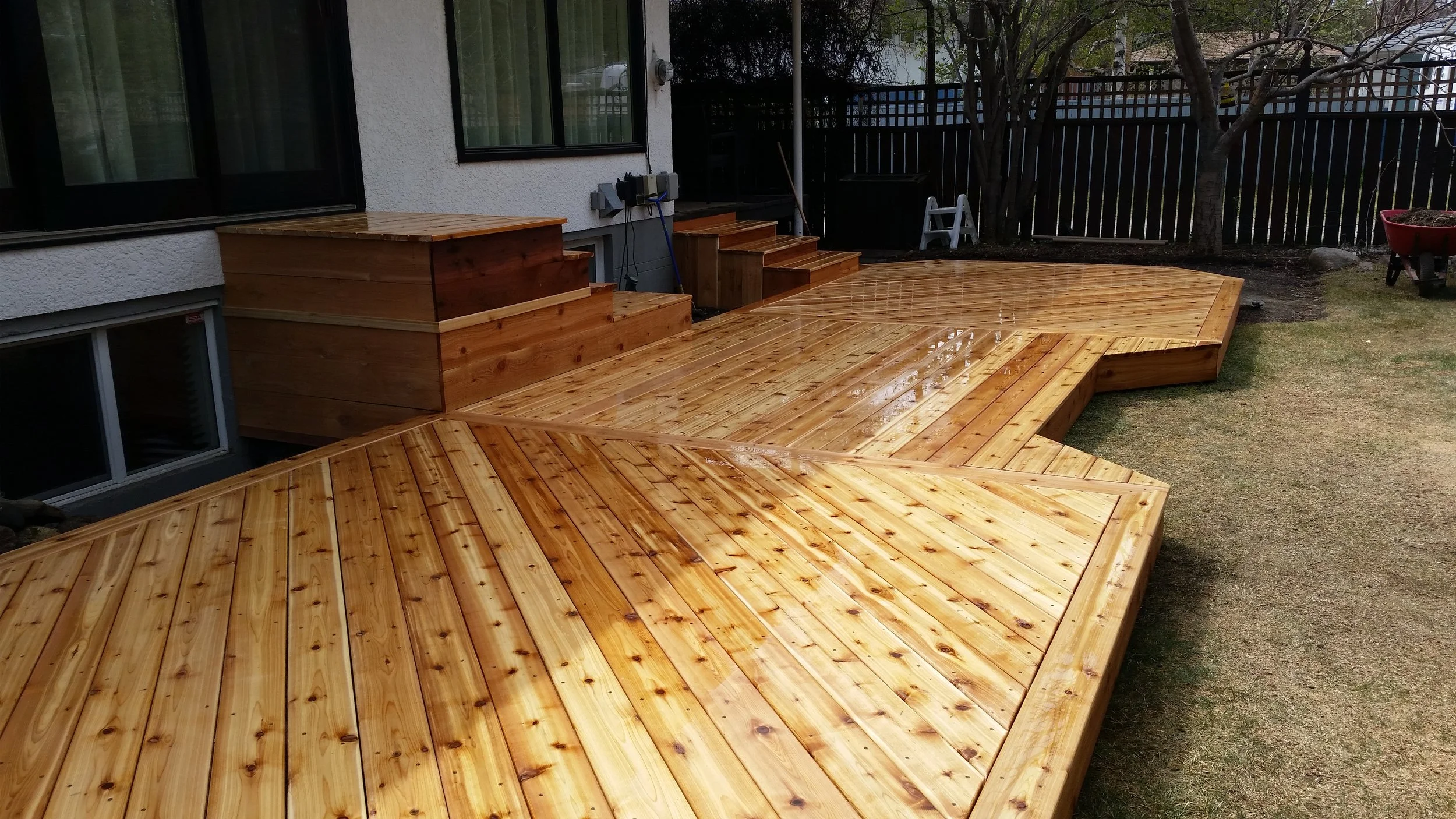 A newly built wooden deck in a backyard, with multiple levels and steps, wet from recent rain, next to a house with sliding glass doors and a white exterior wall.