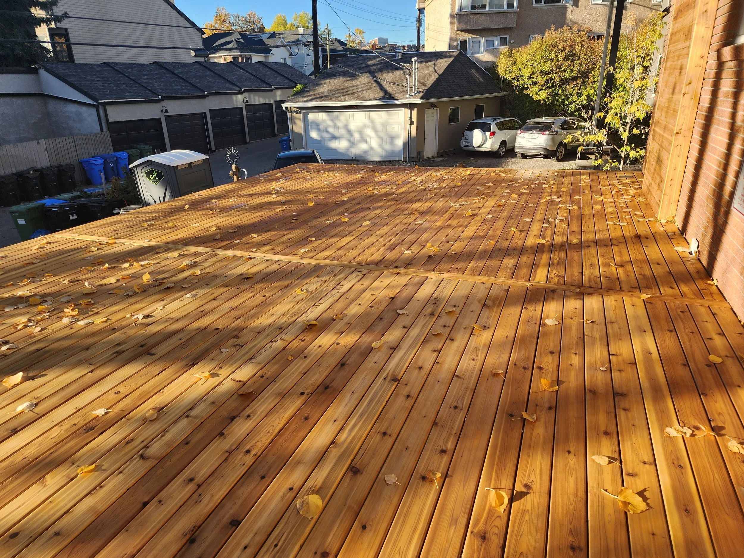View of a backyard with a wooden deck, fallen leaves, parked cars, small building, trees, and neighboring houses and buildings.