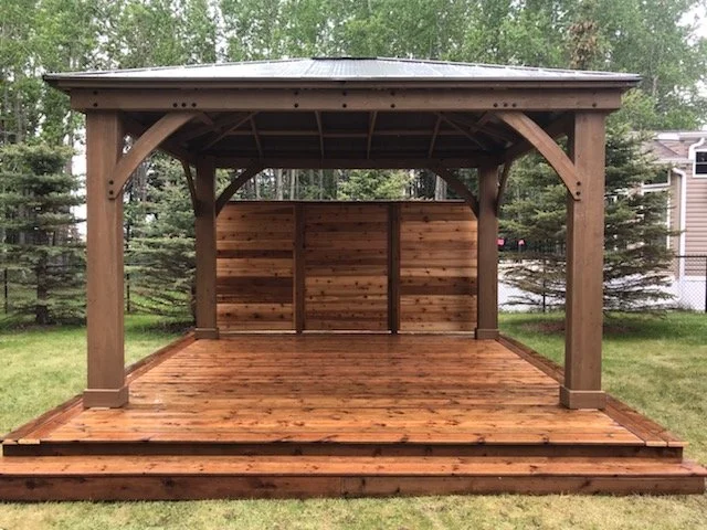 A wooden outdoor gazebo with a roof, built on a deck with steps, surrounded by grass and trees.