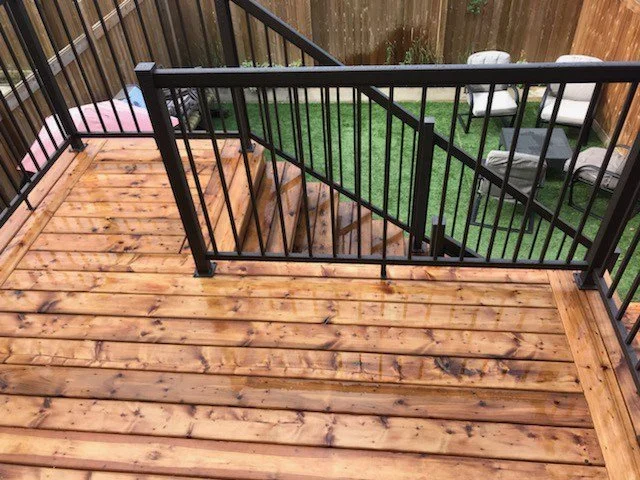 View of a wooden deck with a black metal railing, overlooking a backyard with artificial grass and outdoor chairs.