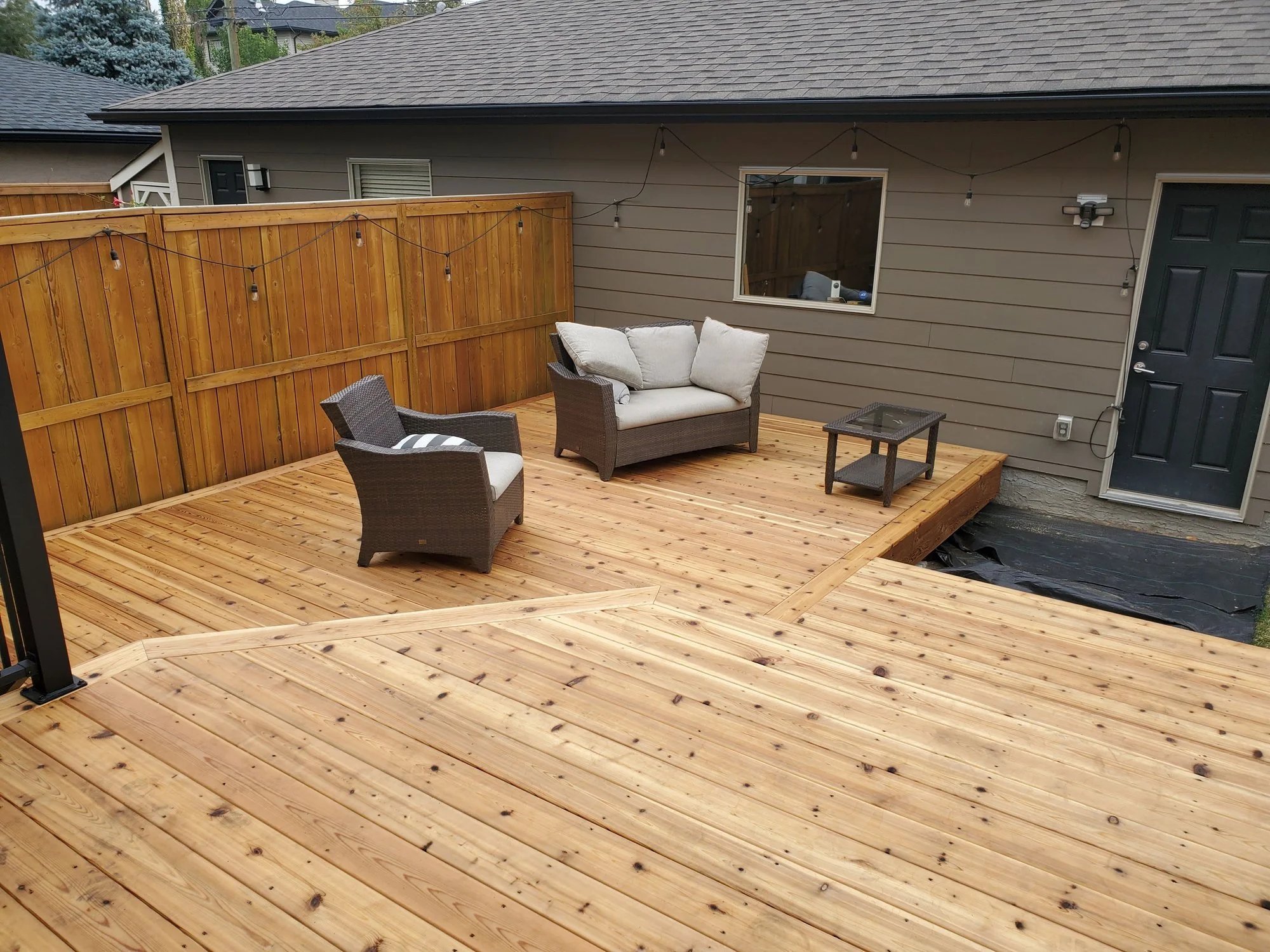 A backyard deck with outdoor furniture, including a loveseat, a chair, and a small table. The deck is enclosed by a wooden fence with string lights, and the house behind has beige siding and a window. Part of the house's roof and neighboring houses are visible.