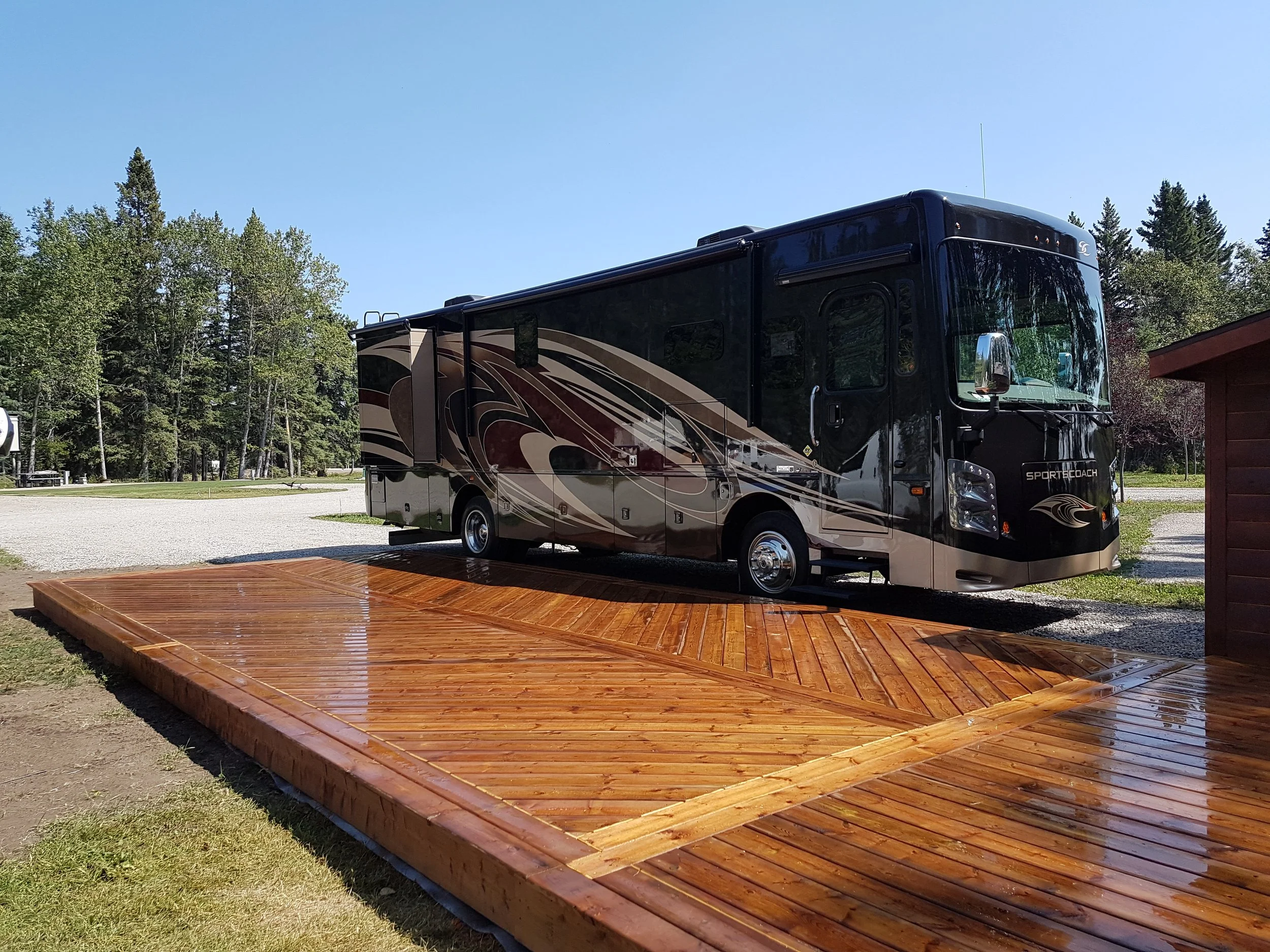 A black and beige recreational vehicle (RV) parked on a gravel lot next to a wooden deck, with green trees and a clear blue sky in the background.