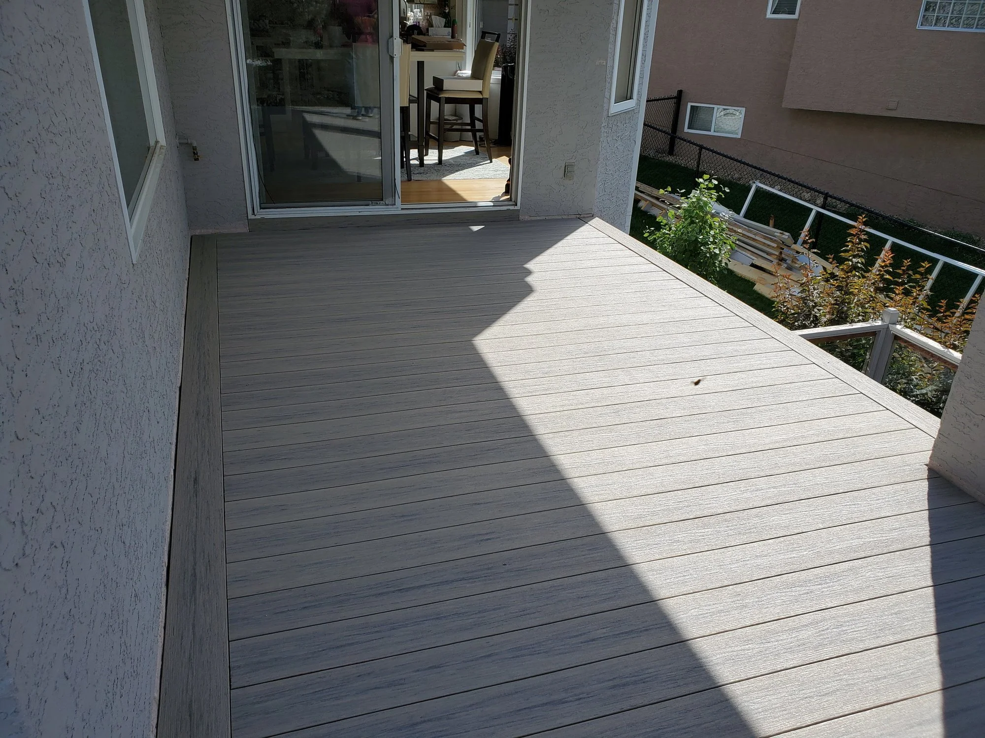 A balcony with light-colored wooden decking, a sliding glass door leading into a room with a dining table and chairs, and a small garden with plants and a fence beyond.