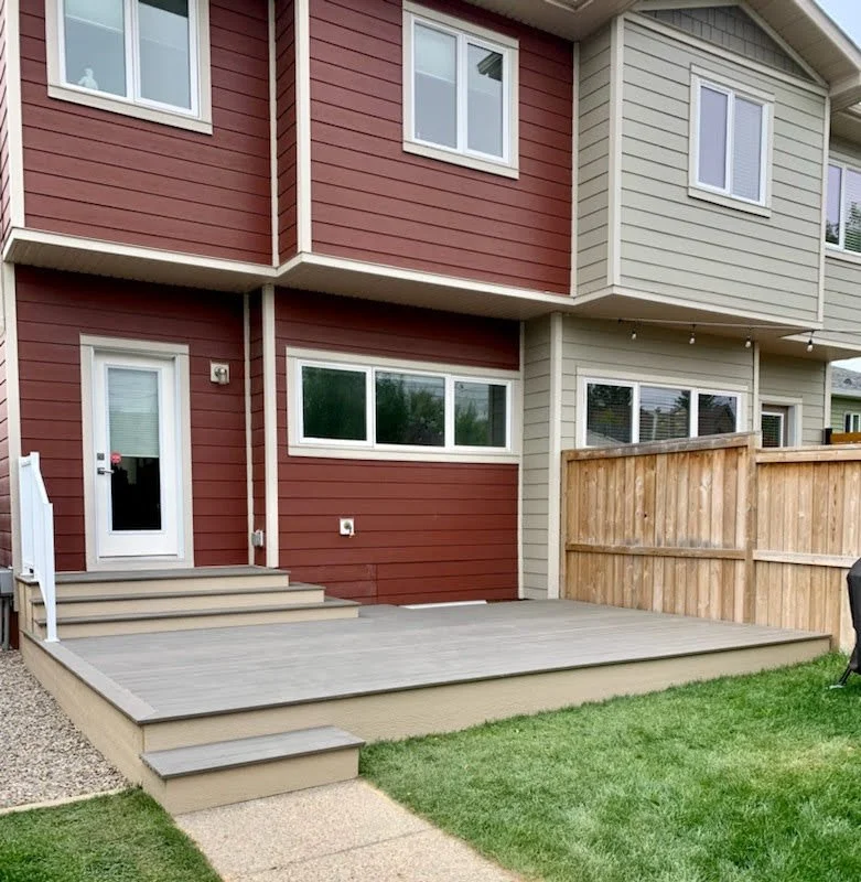 Backyard with a multi-level wooden deck, steps leading up to a door, a green lawn, and a wooden fence. The house has red and beige siding with multiple windows.