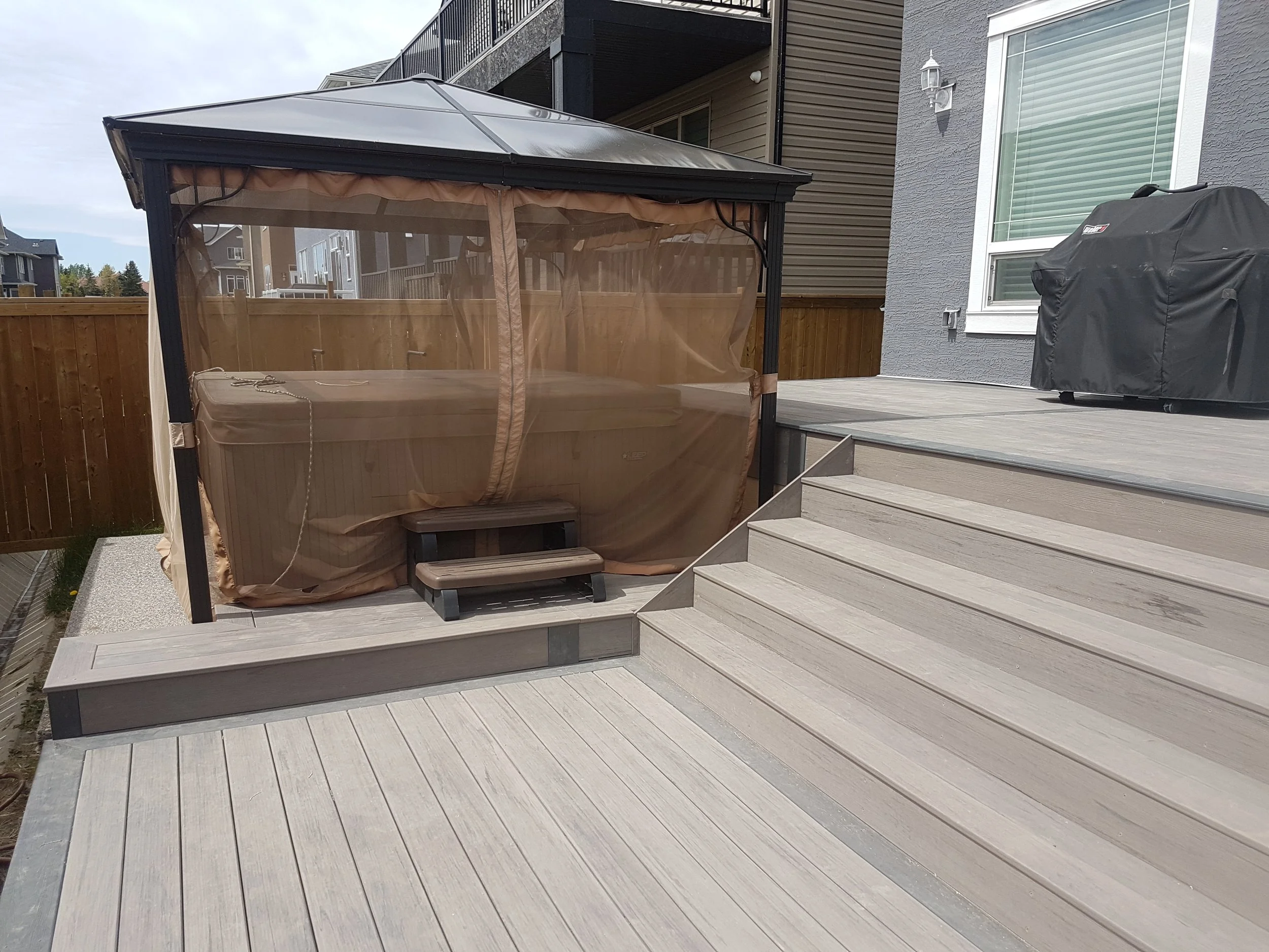 A backyard deck with stairs leading up to a house. There is a covered hot tub and a charcoal grill on the deck. The house has light blue siding and a large window with blinds. A wooden fence surrounds the yard and neighboring houses can be seen in the background.