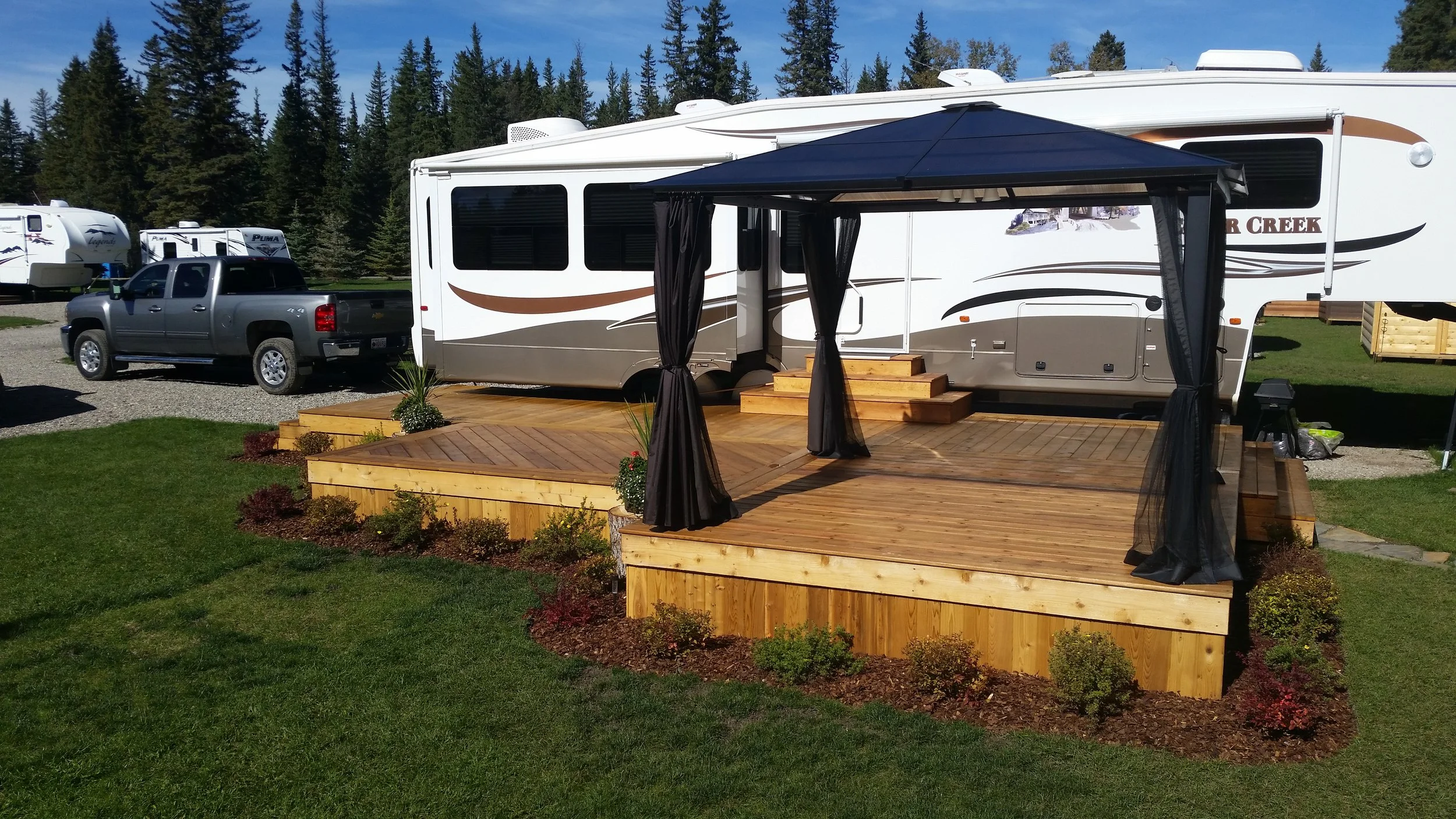 A white travel trailer with brown and black decorative stripes parked on a gravel lot, with a gray pickup truck attached. There is a wooden deck with steps and black curtains set up in front of the trailer, and a small plant in a pot on the deck. In the background are pine trees and other trailers.