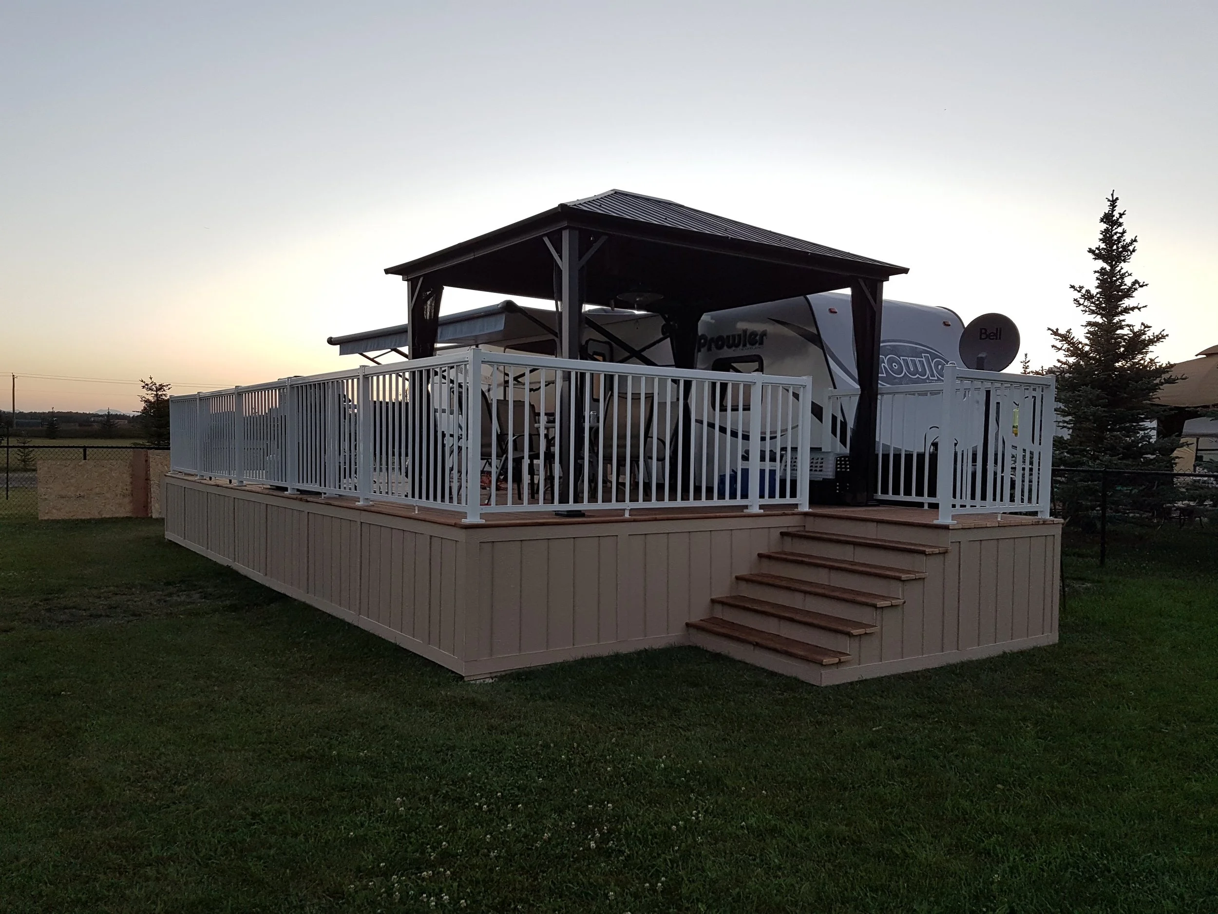 A wooden deck with stairs surrounds a large RV parked underneath a small roofed structure with a metal roof, set in an open grassy area with a few trees and a distant fence at sunset.