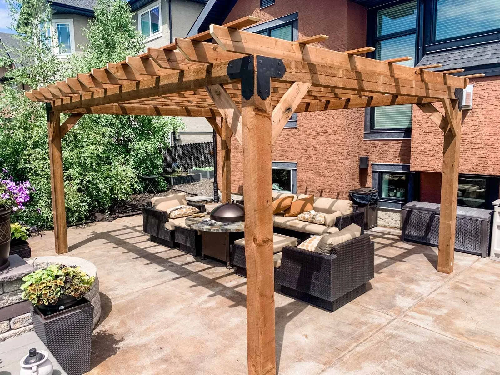 Backyard patio with outdoor seating under a wooden pergola, surrounded by greenery and a brick house.
