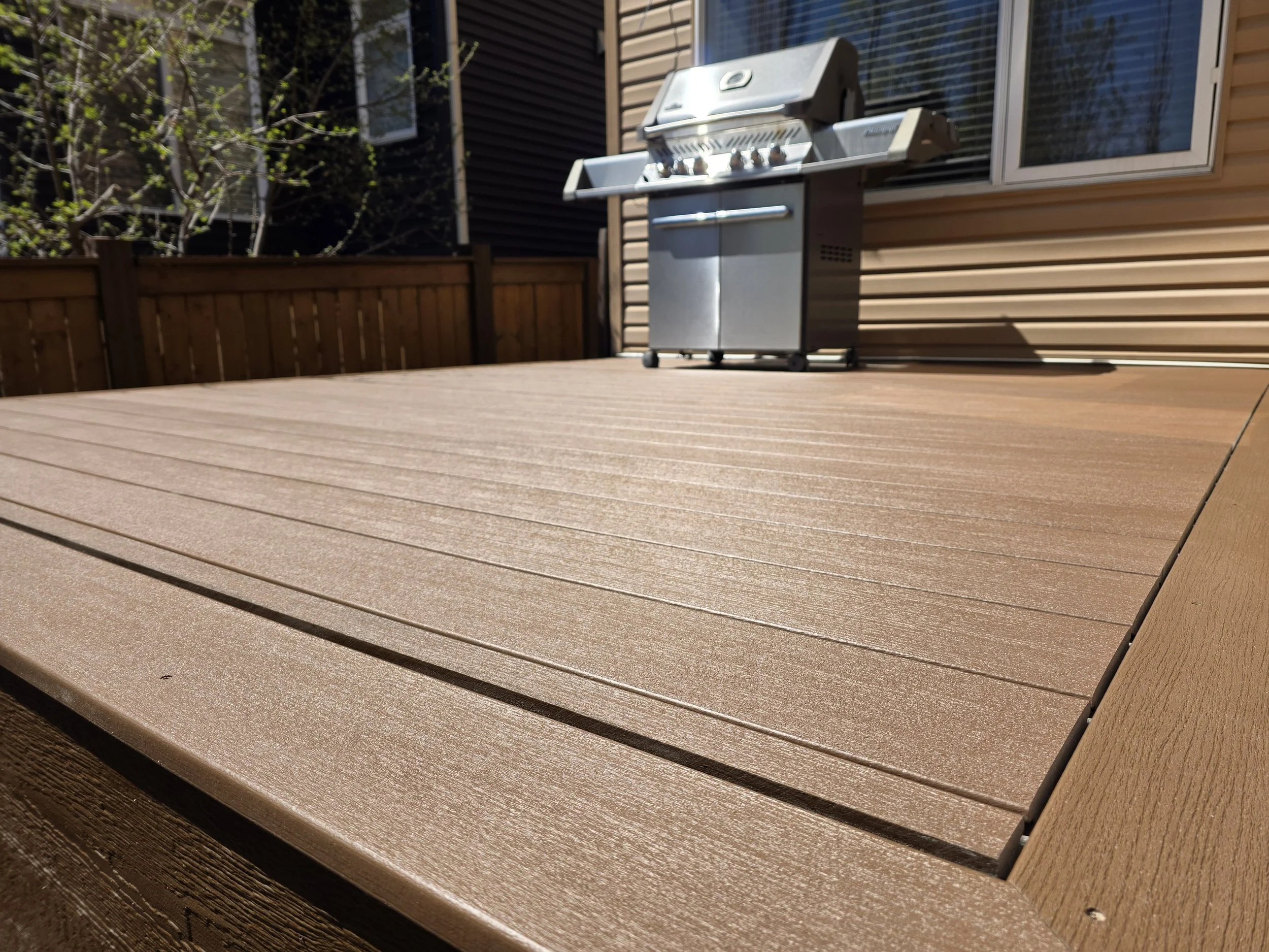 An outdoor wooden deck with a stainless steel gas grill near a house with brown siding and a sliding glass door, surrounded by a wooden fence and trees.