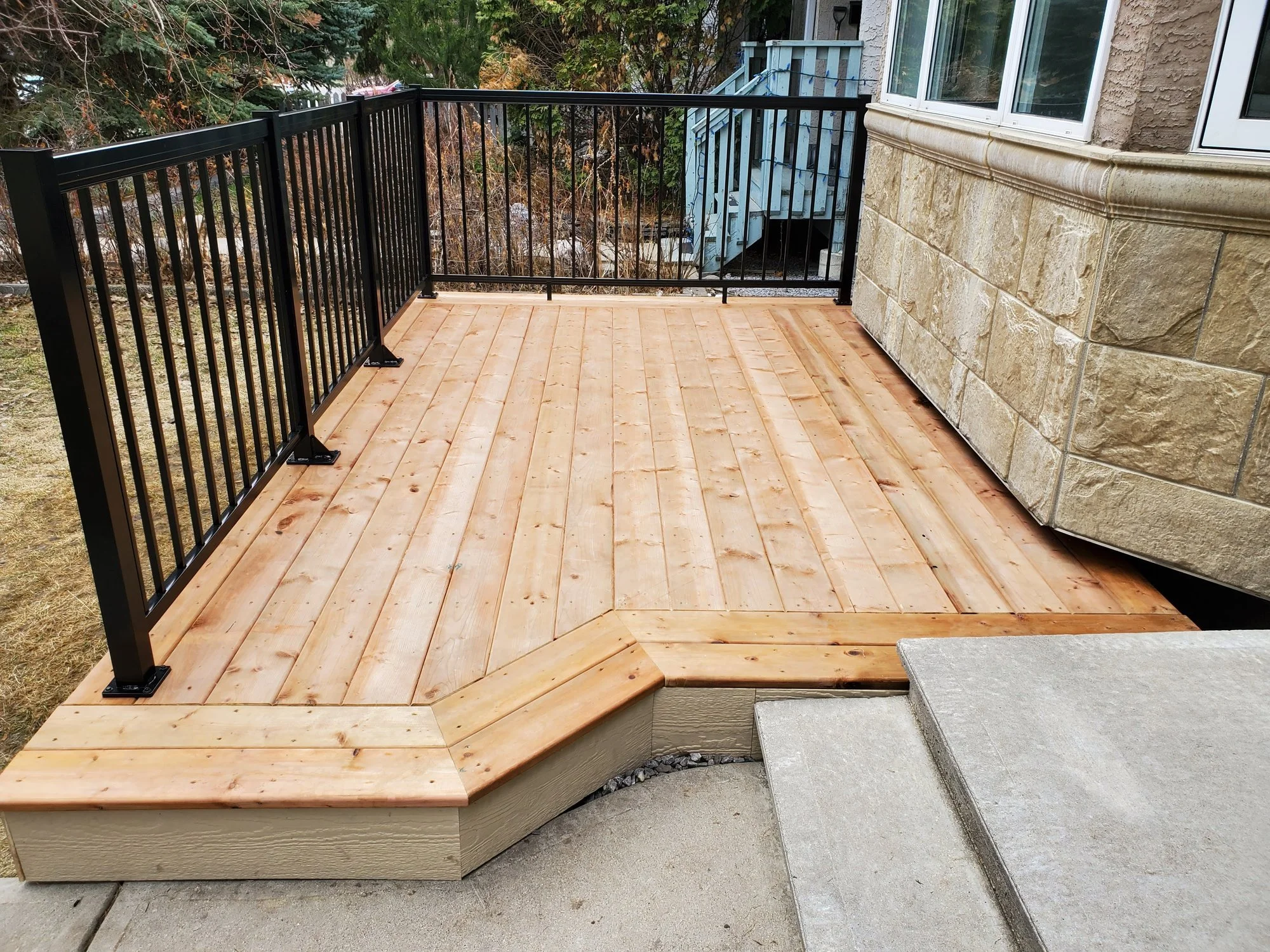 Newly built wooden deck with black metal railing attached to a house with stone exterior walls and a window. The deck has steps leading down to concrete and gravel ground, with trees visible in the background.