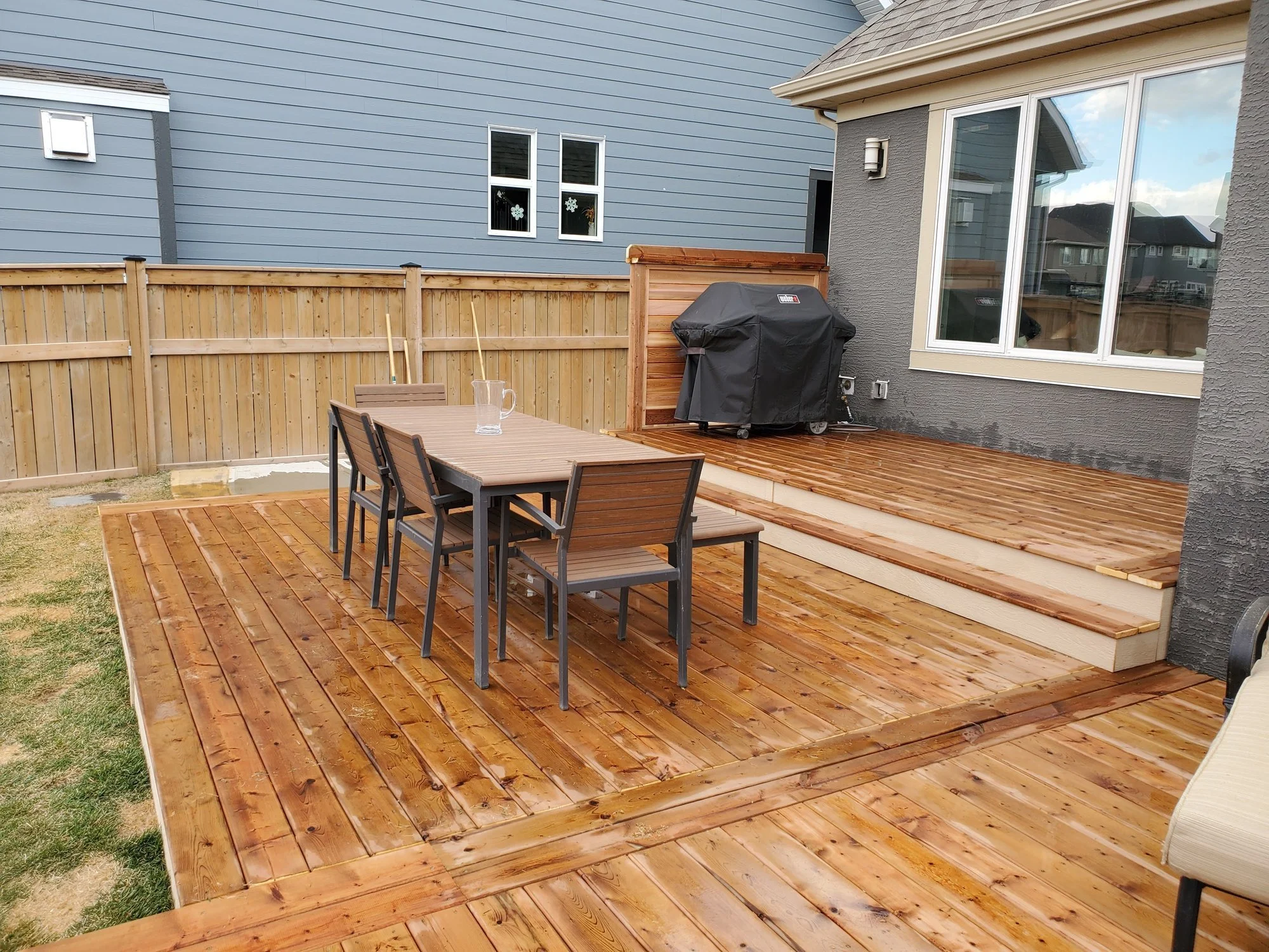 Backyard patio with wooden deck, outdoor dining table with chairs, and a barbecue grill covered with a black cover, fenced with a wooden privacy fence, and neighboring houses visible in the background.