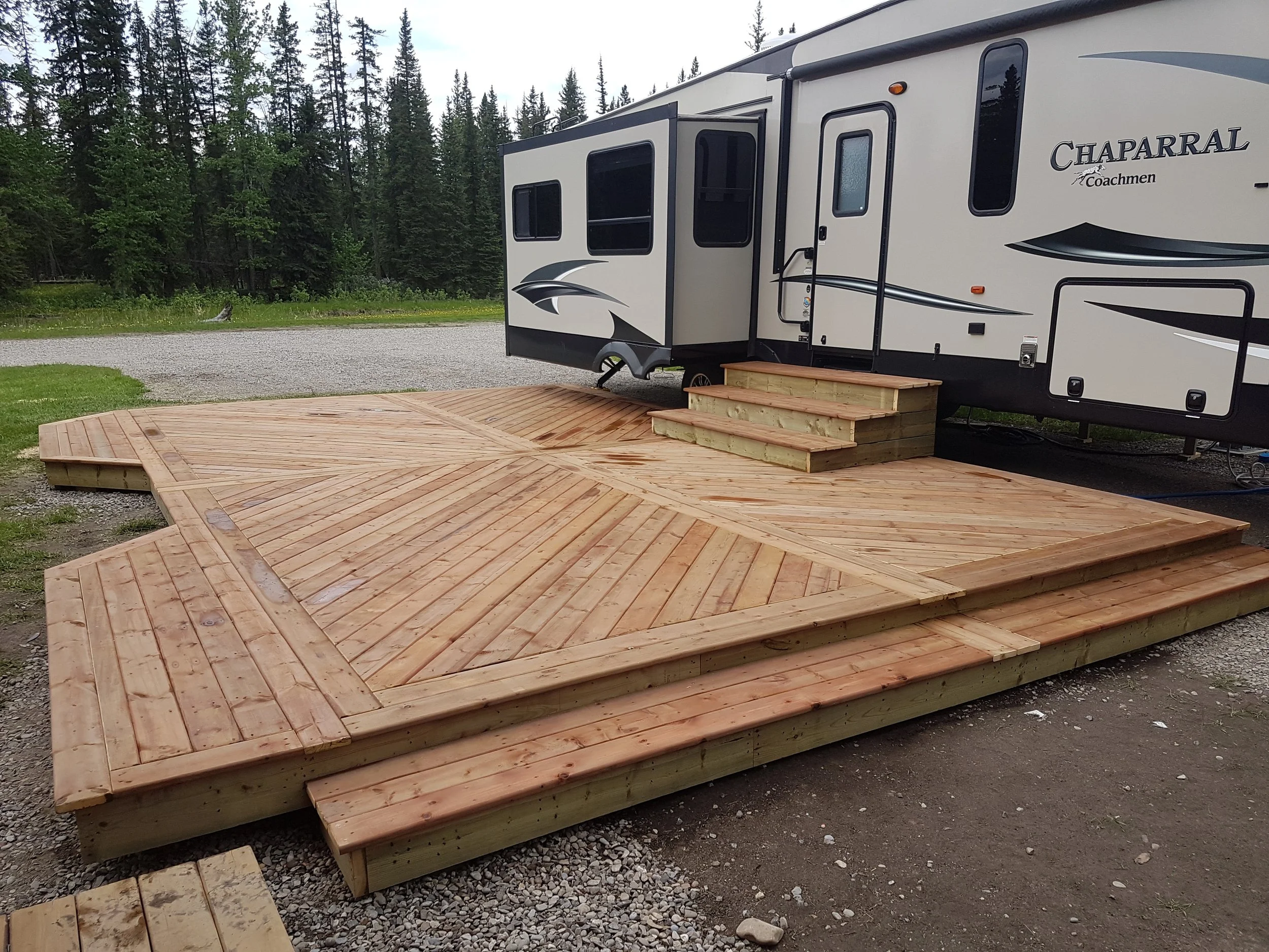A white and black Chaparral Coachmen RV parked on a gravel area next to a forest of tall pine trees. In front of the RV, there is a newly built wooden deck with steps.