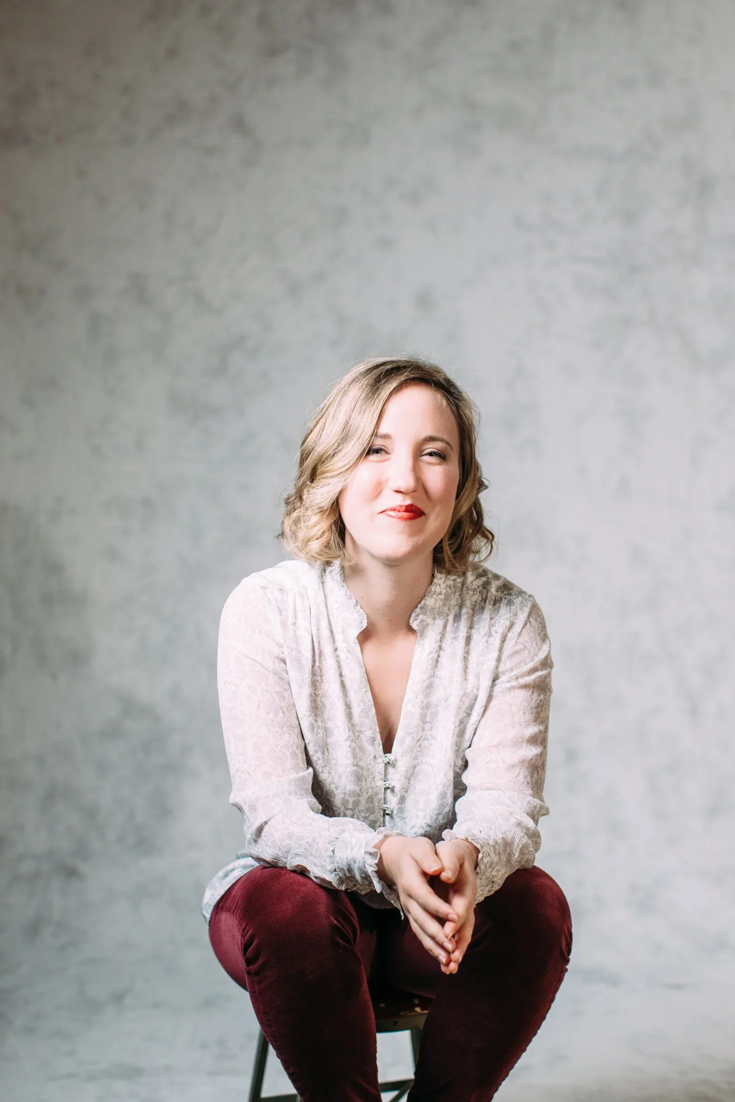 A woman with short blond hair, wearing a light-colored blouse, sitting against a neutral background, smiling with one eye slightly closed.