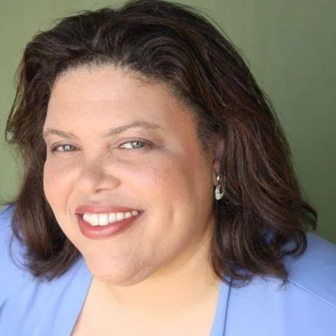 Close-up portrait of a smiling woman with curly dark hair, red lipstick, and earrings, wearing a blue outfit.