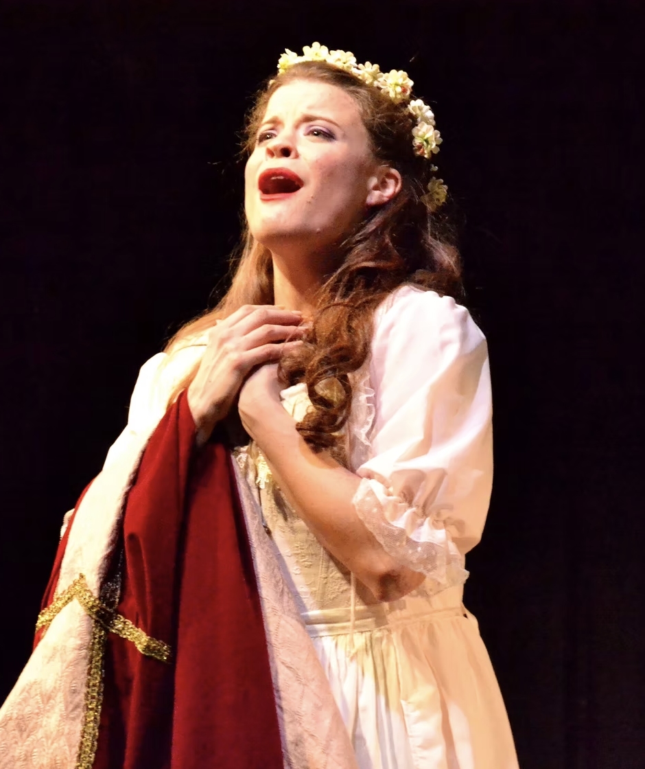 A woman with wavy, shoulder-length hair, wearing a red sleeveless top, smiling with red lipstick, posing with her hand touching her hair, and a quote overlay Reads: ‘Studying with Dana has been and continues to be transformative.’ - Tamara Ryan, Soprano.