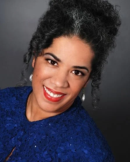 Close-up portrait of a smiling woman with curly dark hair, red lipstick, and earrings, wearing a blue outfit.