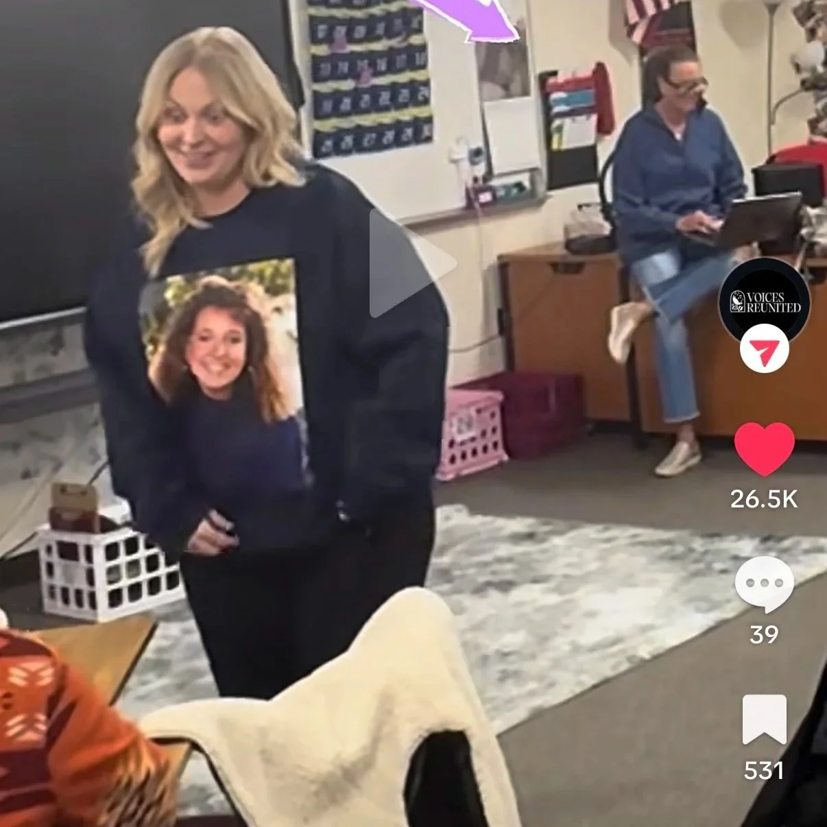 A woman with blonde hair wearing a black sweatshirt with a large photo of herself, smiling, inside a classroom or office. In the background, a woman in a blue jacket seated on a desk, working on a laptop. The room has a whiteboard, posters, and storage bins.