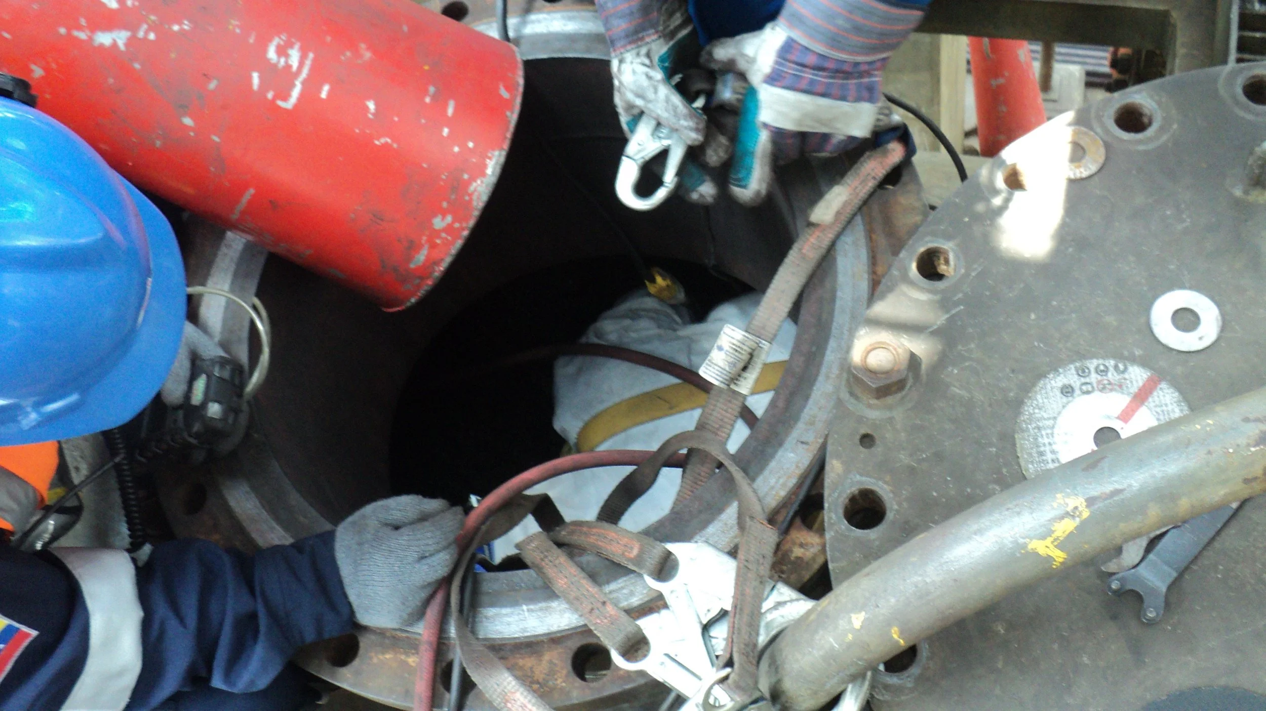 Trabajador en una instalación industrial, usando casco azul y guantes, realiza tareas en una estructura de metal con herramientas y equipos de seguridad, incluyendo un cilindro de gas y cinturones de seguridad.