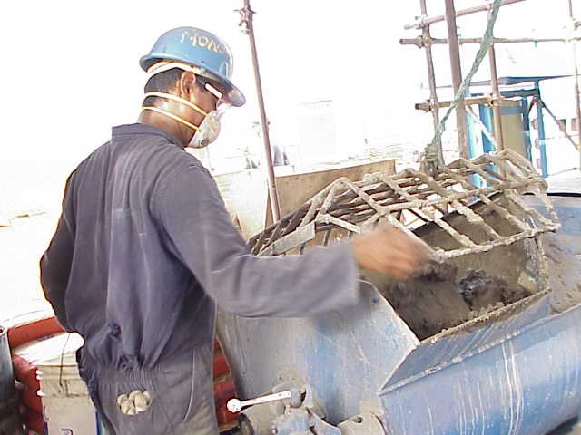 Trabajador en construcción, usando casco, máscara y lentes de protección, manejando mezcladora de cemento.