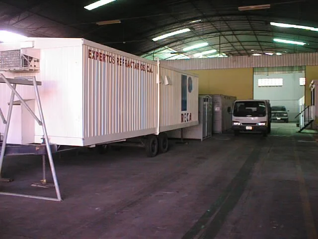 trailer móvil blanca con letras rojas que dicen 'Expertos Refractarios CA', en un espacio cubierto con vehículos estacionados al fondo.