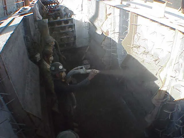 Dos trabajadores en equipo usando casco, gafas y máscara de protección, trabajando en una Estructura de acero con anclajes refractarios en un sitio de construcción.