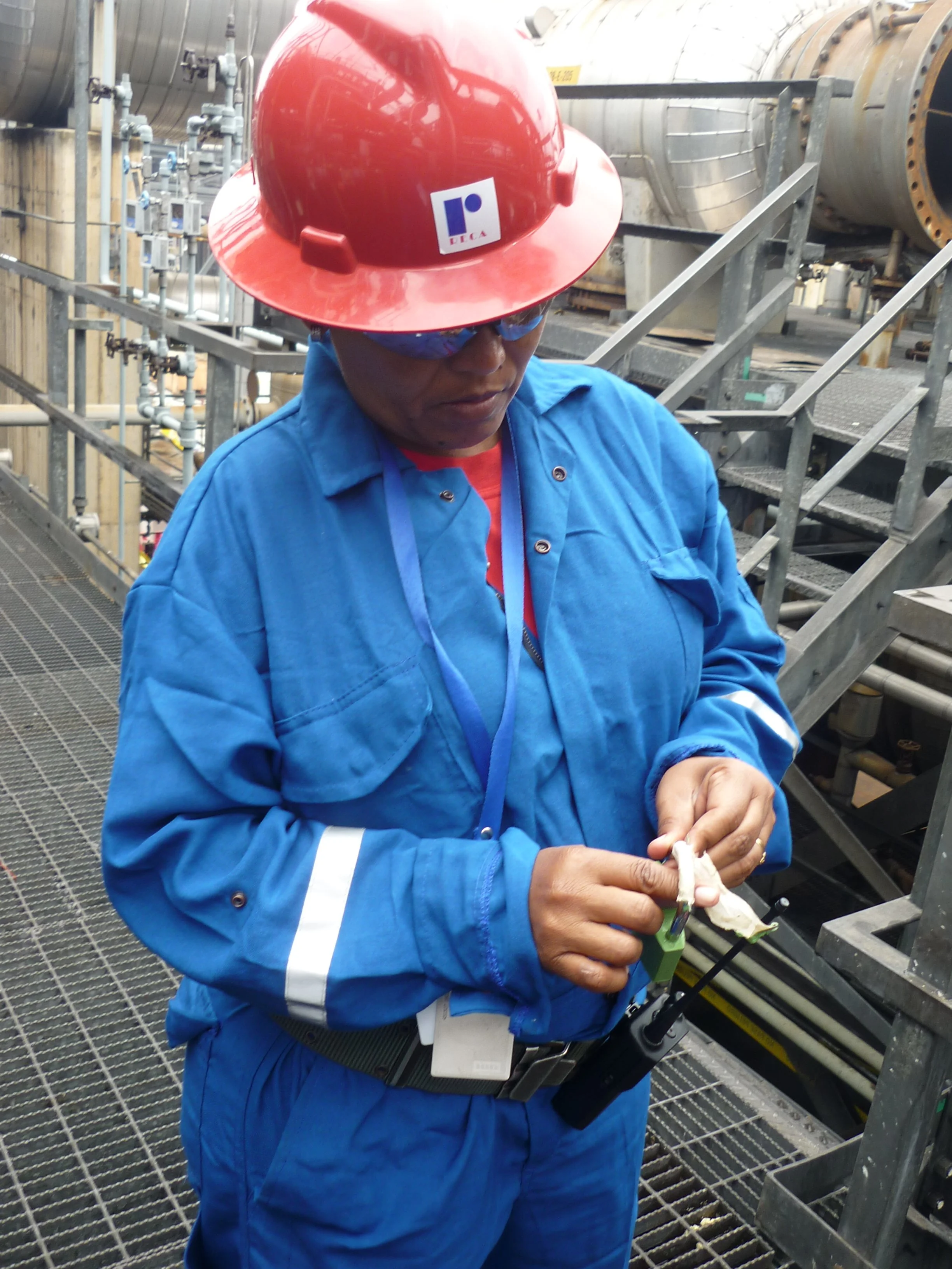Operaria en un entorno industrial revisando un dispositivo de seguridad en una planta de manufactura, viste casco rojo, gafas de protección y ropa de trabajo azul.