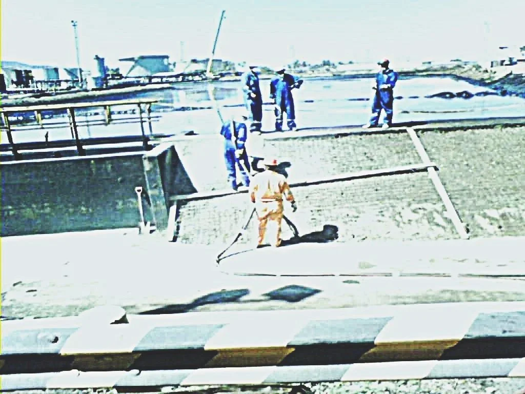 Varios trabajadores construyen en una obra, algunos con equipo de protección, en un sitio con agua y estructura de cemento.