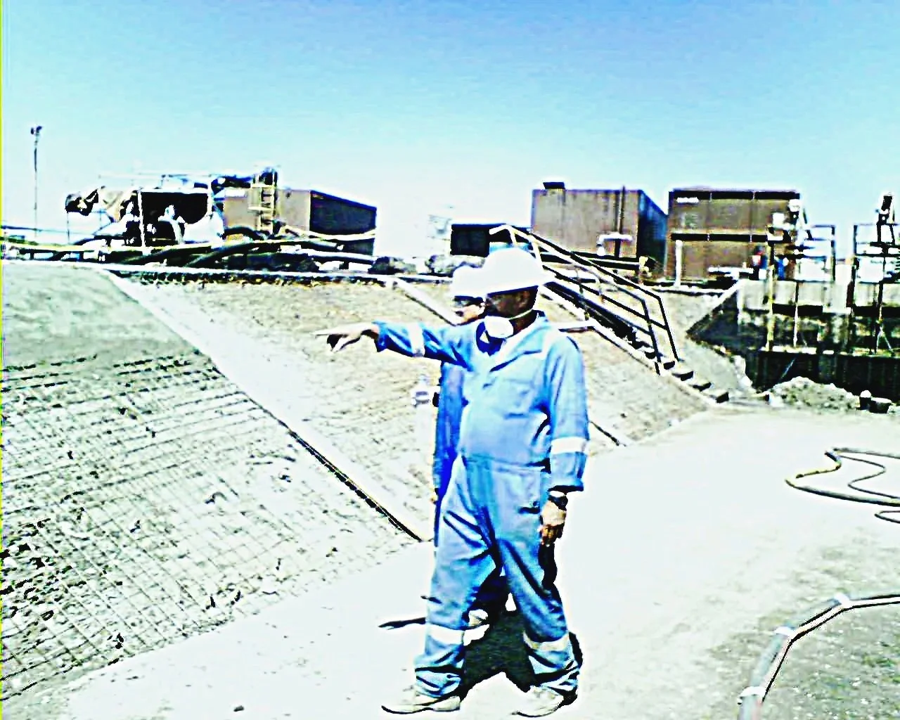 Ingeniero o trabajador de la construcción con casco y mascarilla, señalando hacia un área de construcción en una obra en curso al aire libre.