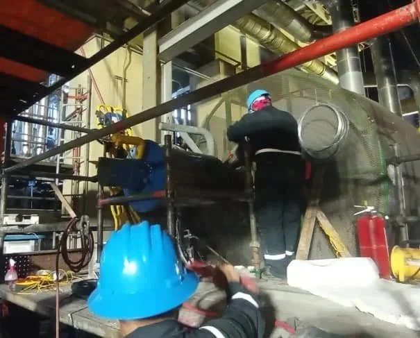Dos trabajadores en un ambiente industrial manipulando maquinaria grande, uno con casco azul y otro con casco. Persona con casco en una planta industrial operando equipo portatil de secado dryout