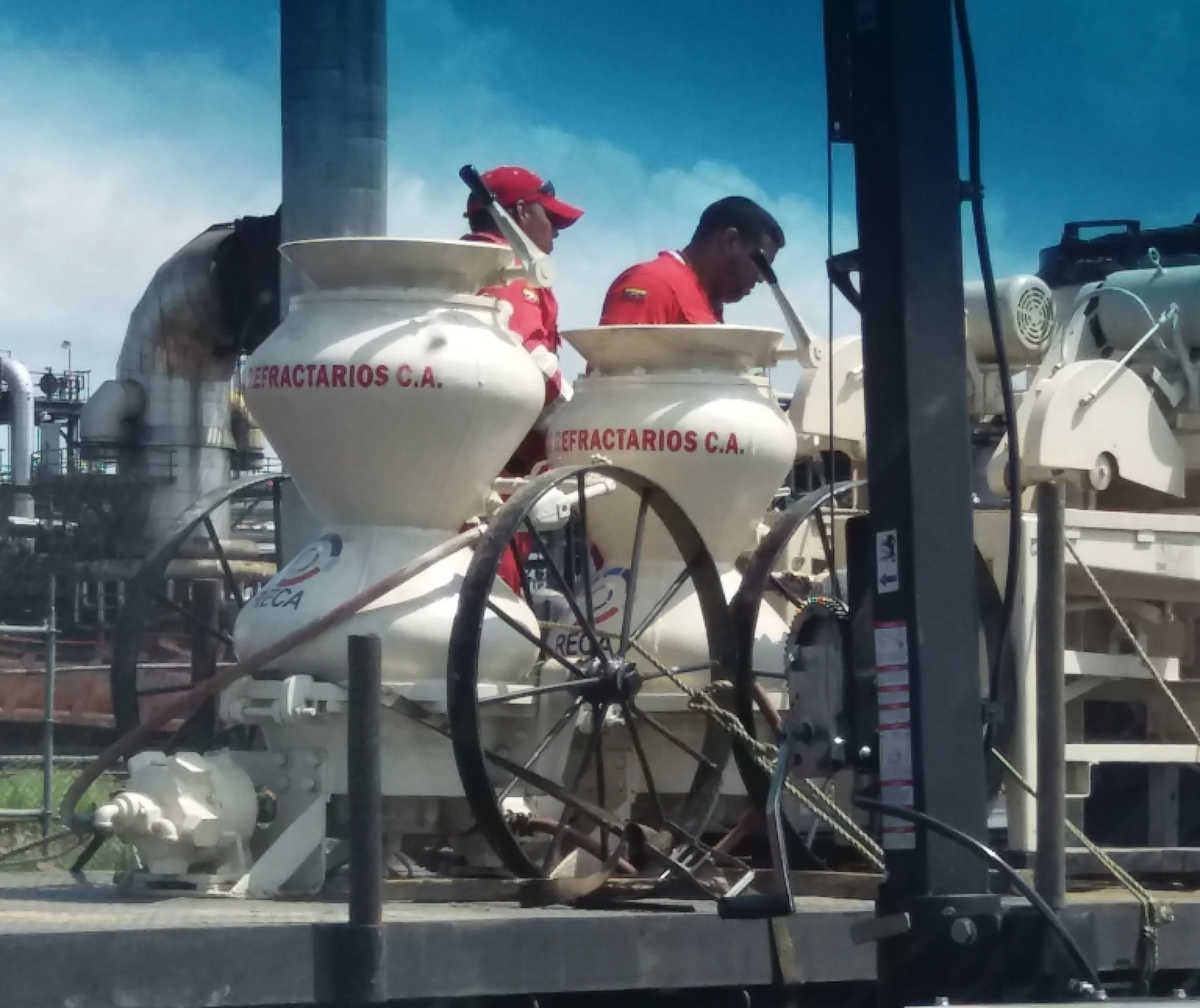 Tres trabajadores en un equipo de construcción en una zona industrial con maquinaria y estructuras metálicas, bajo un cielo despejado.