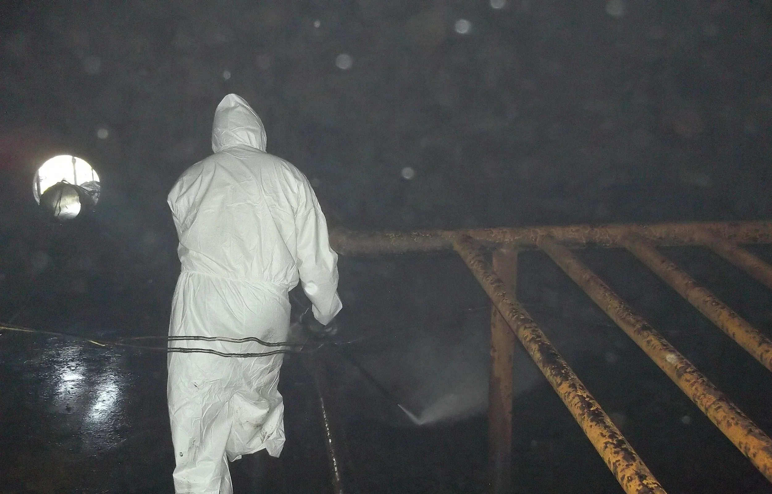 Limpieza quimica. Persona con equipo de protección blanca rociando una superficie en un ambiente oscuro y húmedo con barro barandillas oxidadas.