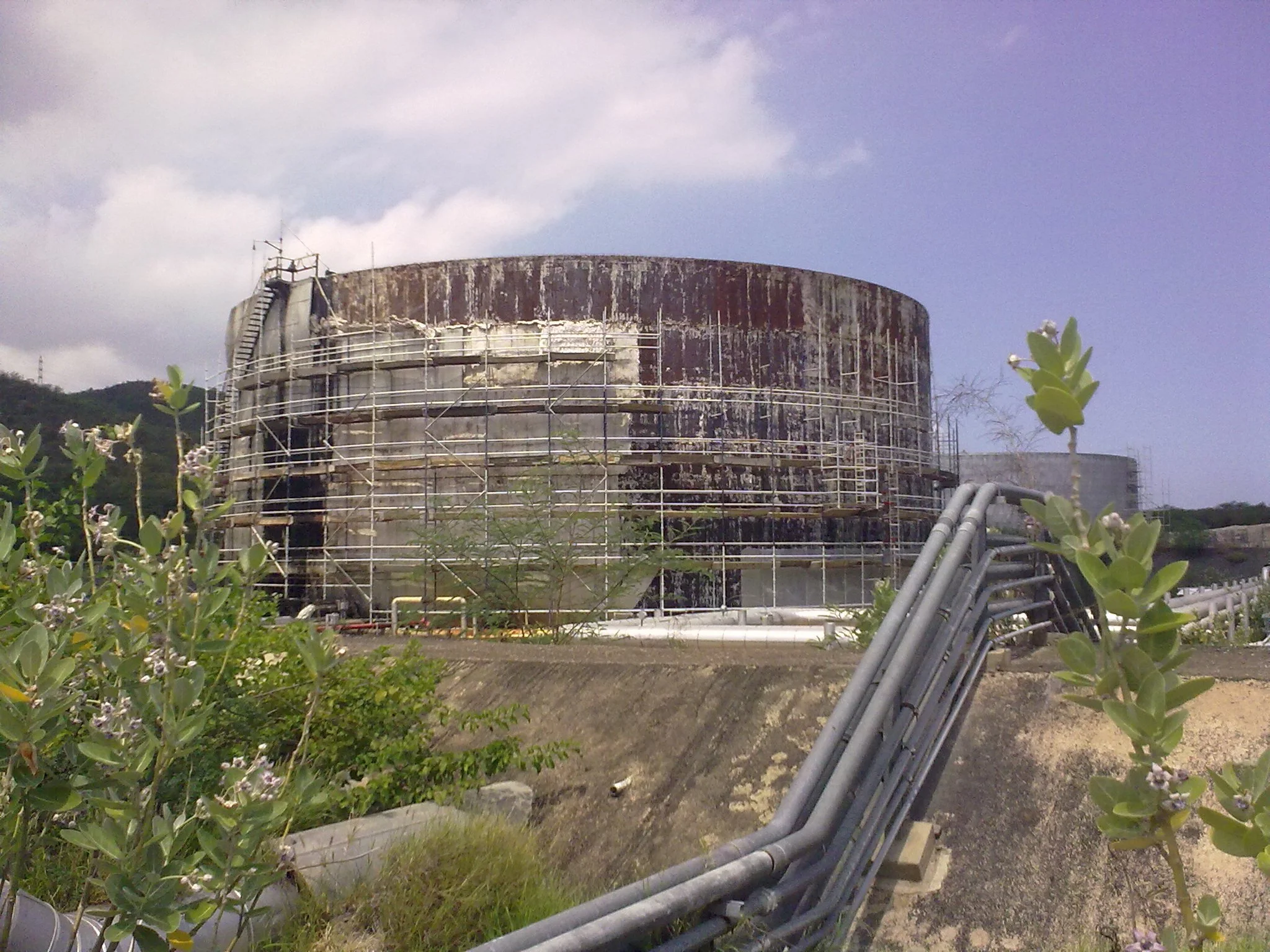 Tanque de almacenamiento  envejecido, rodeado de andamios y tuberías, con vegetación y cielo nublado en el fondo.
