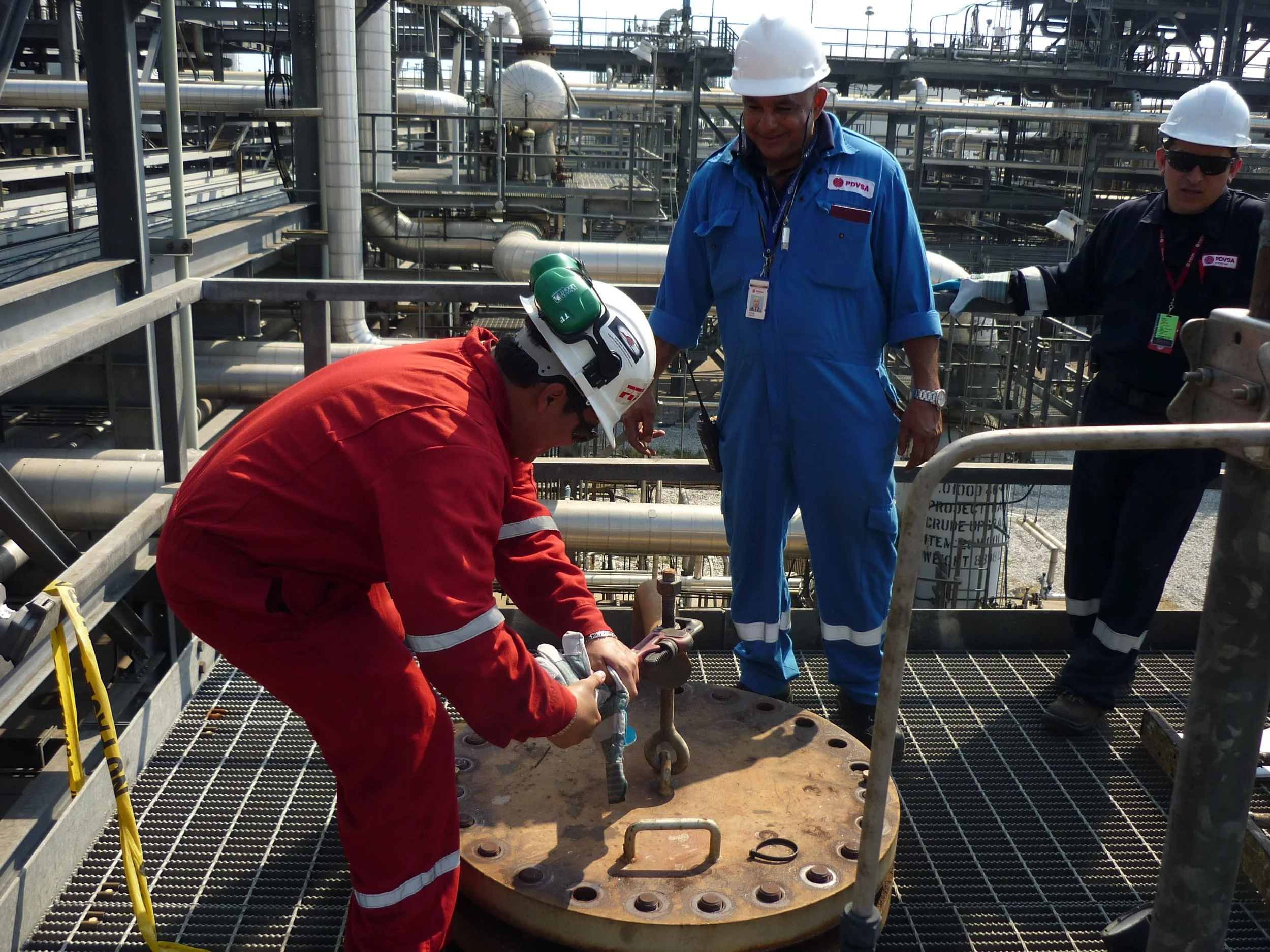 Técnicos trabajando en una plataforma industrial con tuberías y maquinaria, uno en posición agachada manipulando una pieza grande, los otros observando, todos con casco de seguridad y ropa de protección.