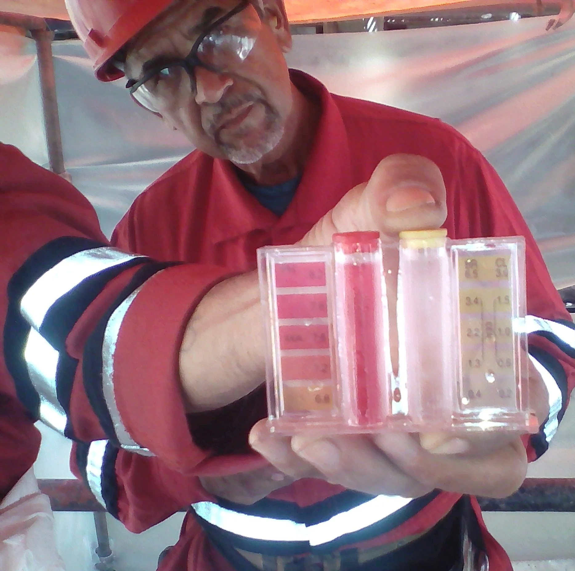 Un trabajador en uniforme , con casco y gafas, sosteniendo un tubo de muestra de color rosa, con instrumentos de medición